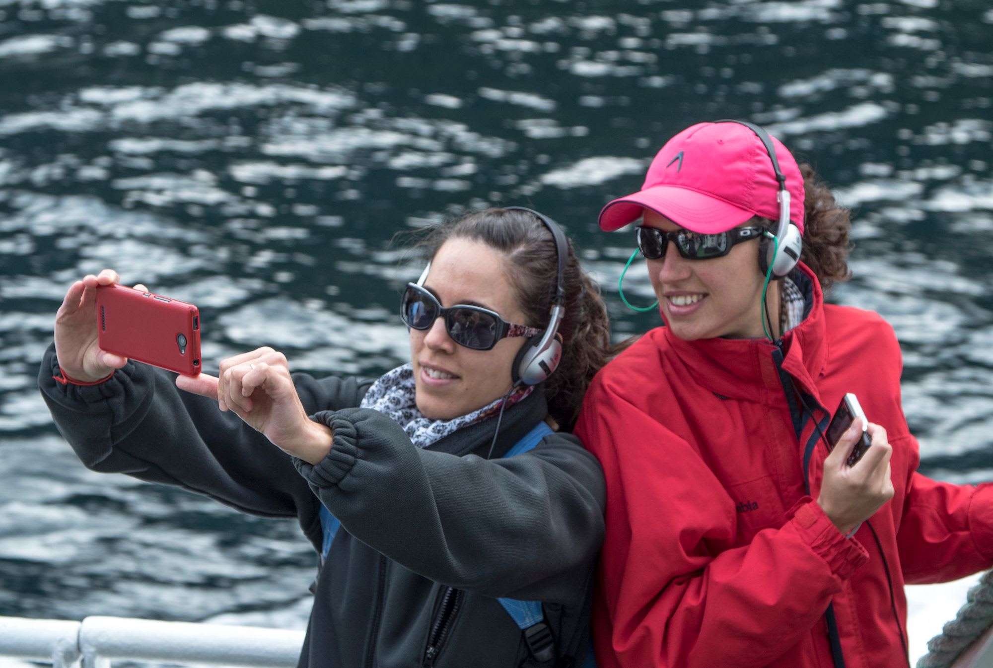 Women in headphones and sunglasses take boat selfie by rippling water.