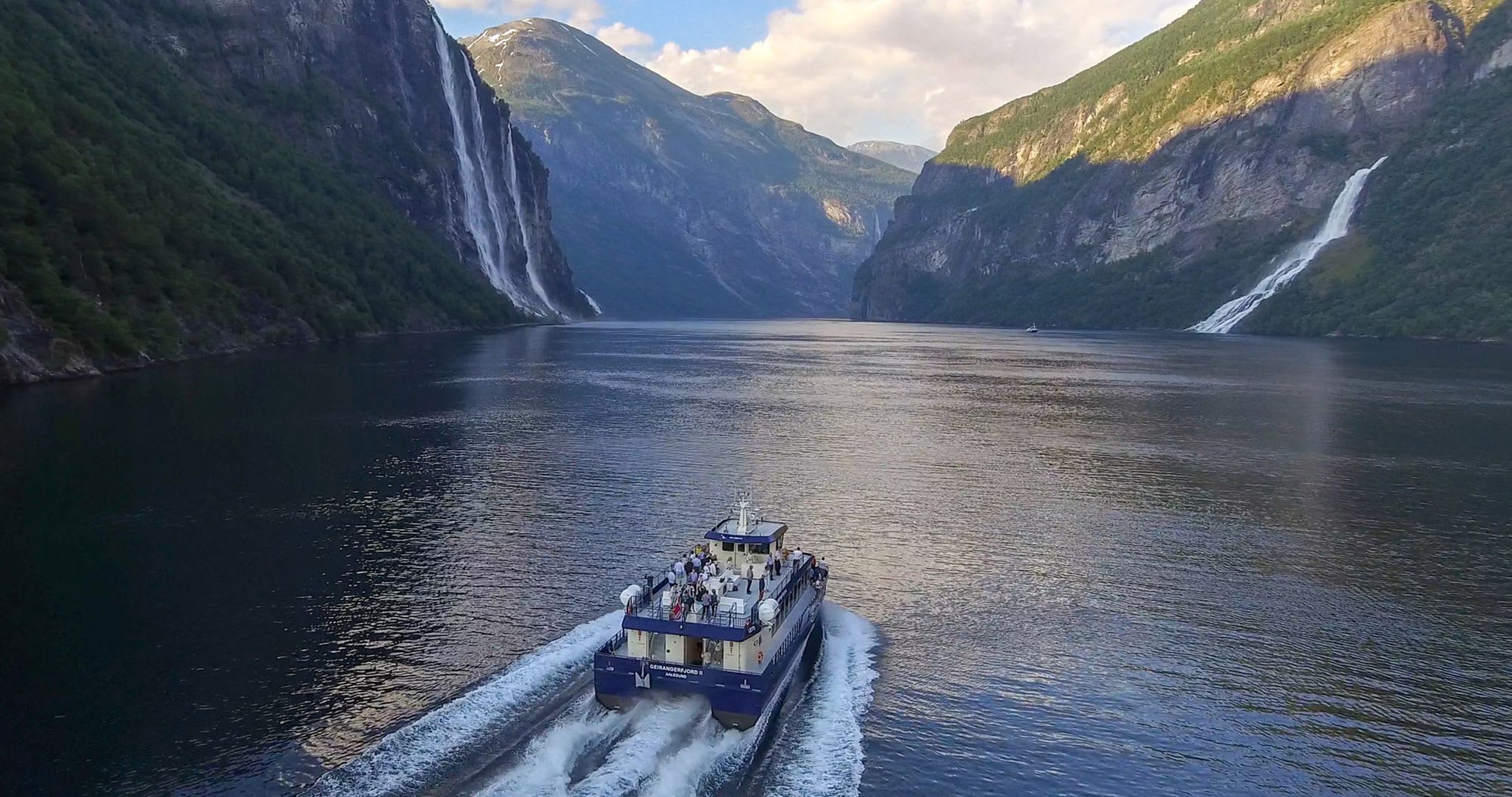 Boot fährt durch engen Fjord mit üppigen Klippen, Wasserfällen und ruhigem, blauem Wasser, das die Landschaft spiegelt.