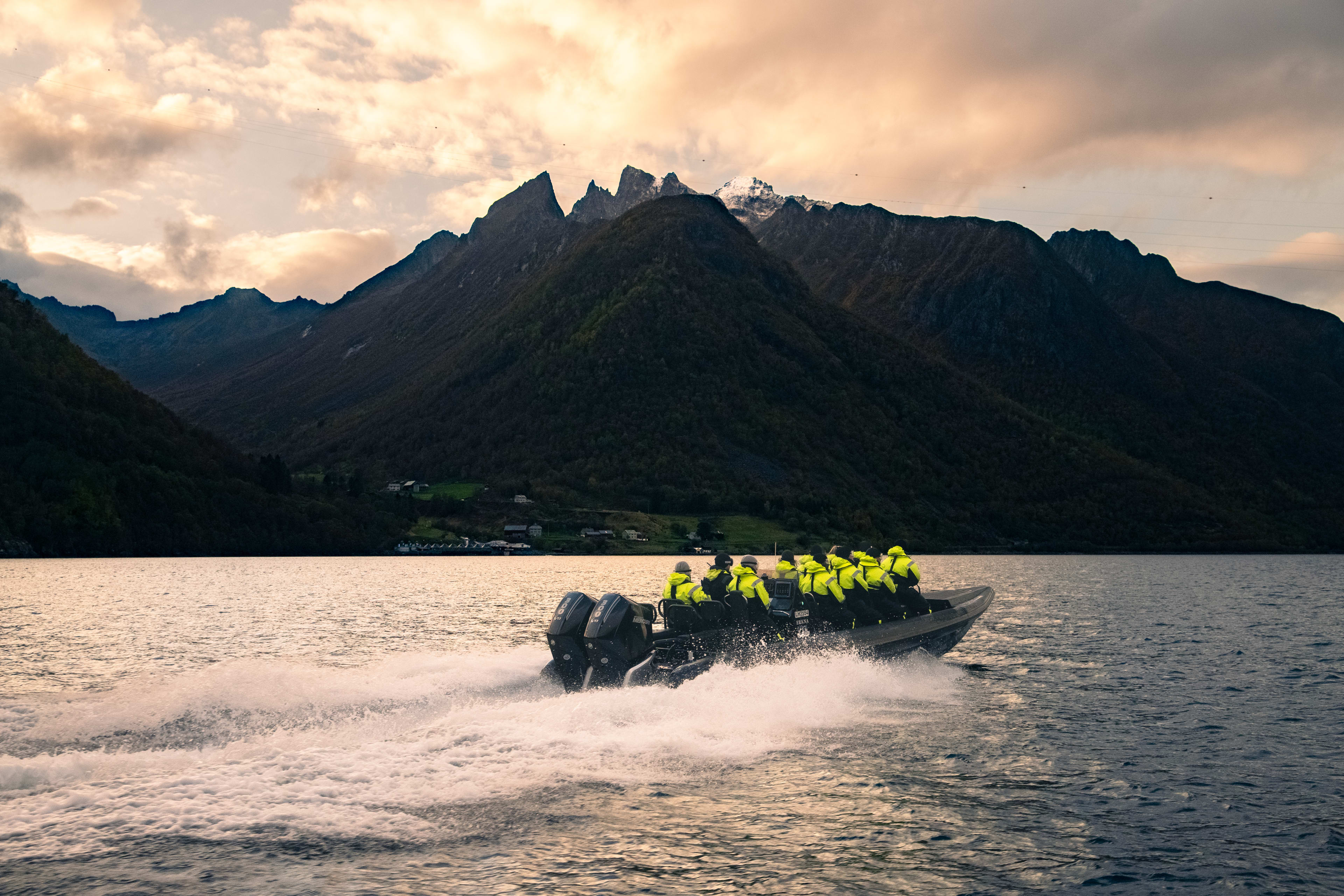 Hurtig RIB-båt gjennom bølger mot Hjørundfjorden fra Ålesund ved solnedgang med dramatiske fjelltopper og fjordlandskap