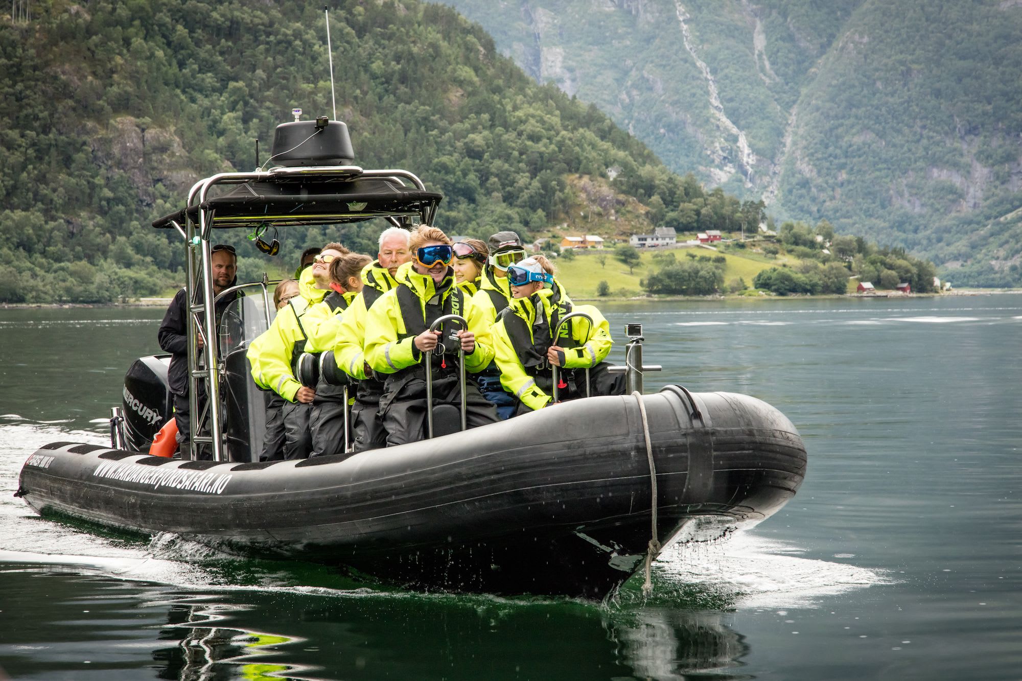 Tourists in yellow ride a boat through fjord with green hills and village.