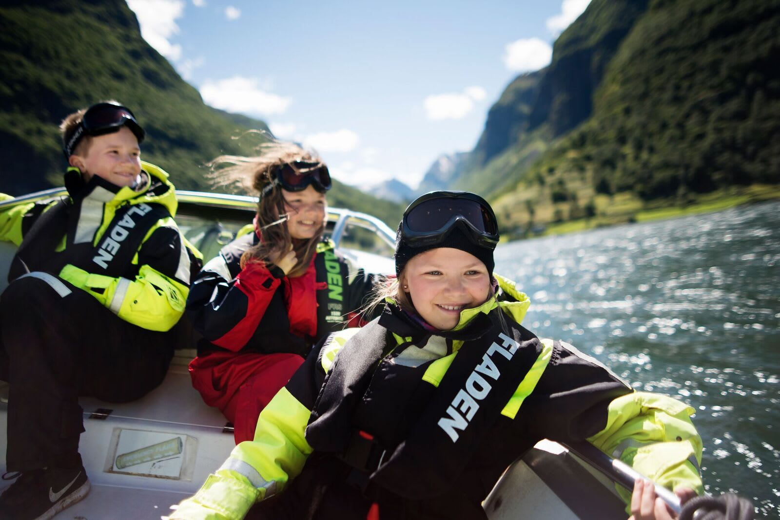 Three smiling kids in waterproof suits ride boat through sunny fjord.