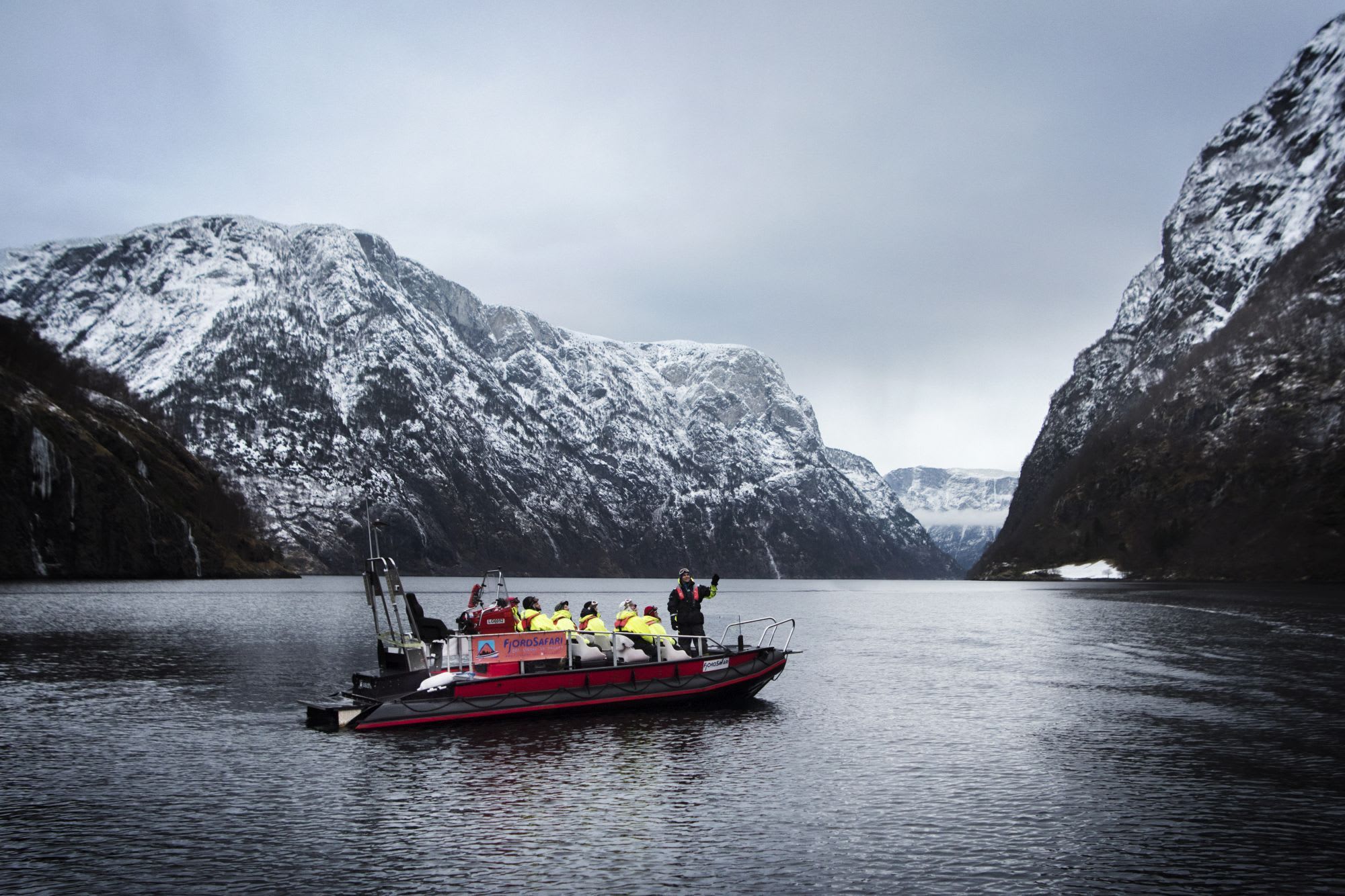 Rødt oppblåsbar båt med personer i gule jakker som flyter på rolig vann, med én person som gestikulerer mot noe.