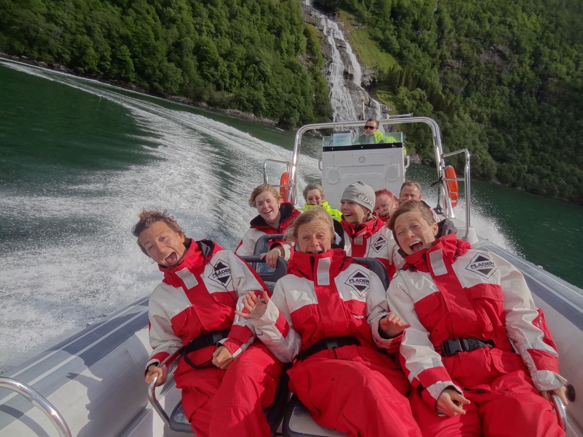 People in red and white waterproof suits on speeding boat, laughing and holding on, leaving a foamy wake.