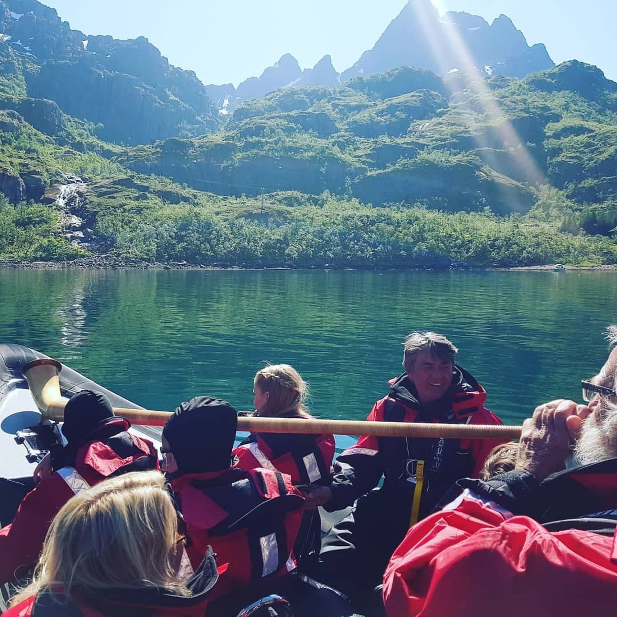 People in red and black waterproof suits on RIB in calm water, surrounded by lush hills and small waterfall in the background.