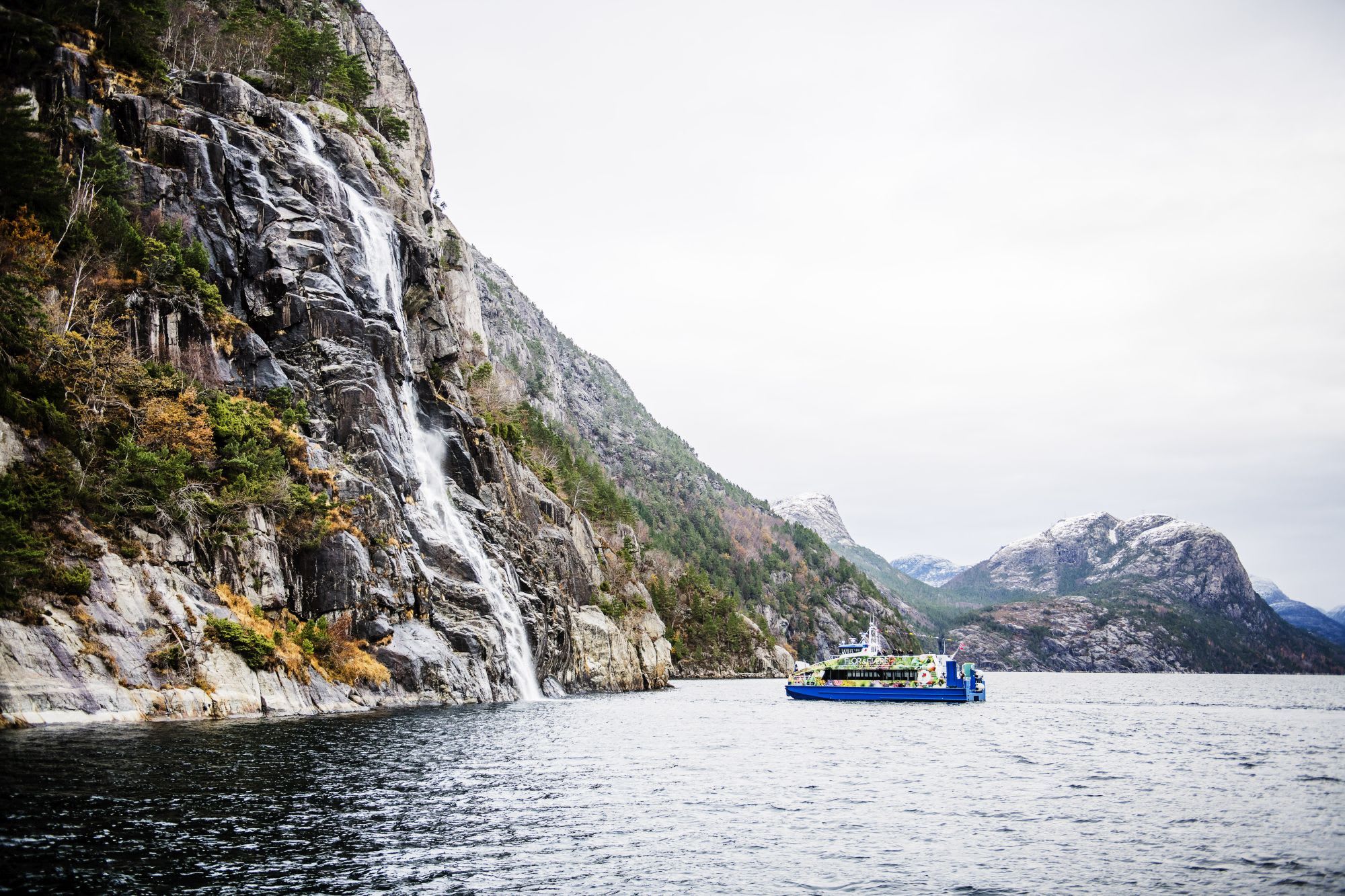 Sightseeing boat near the Hengjanefossen waterfall in the Lysefjord with snowy, forested mountains.