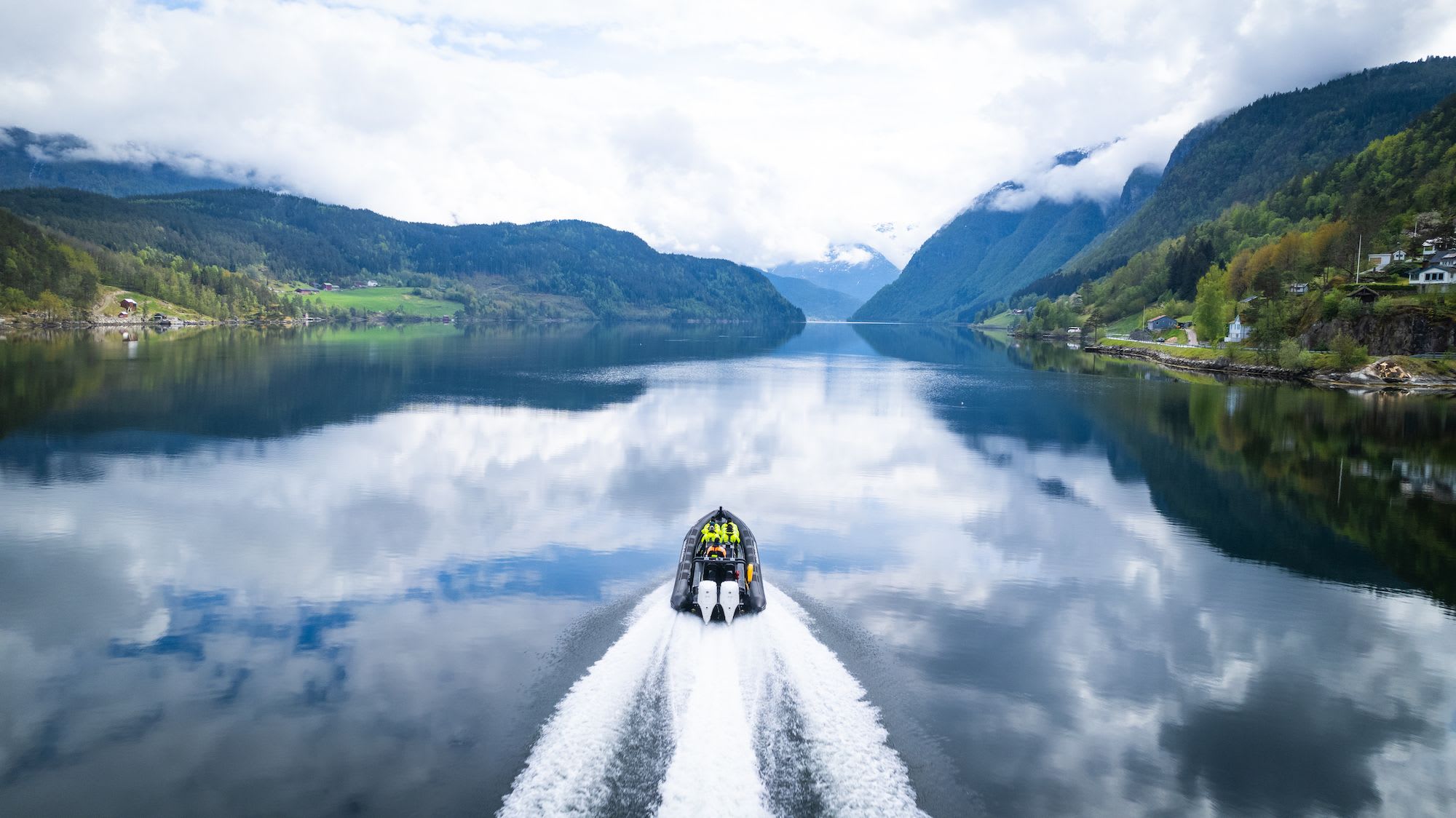 Båt med passasjerer suser over speilblank Hardangerfjord som reflekterer fjell og skyet himmel.