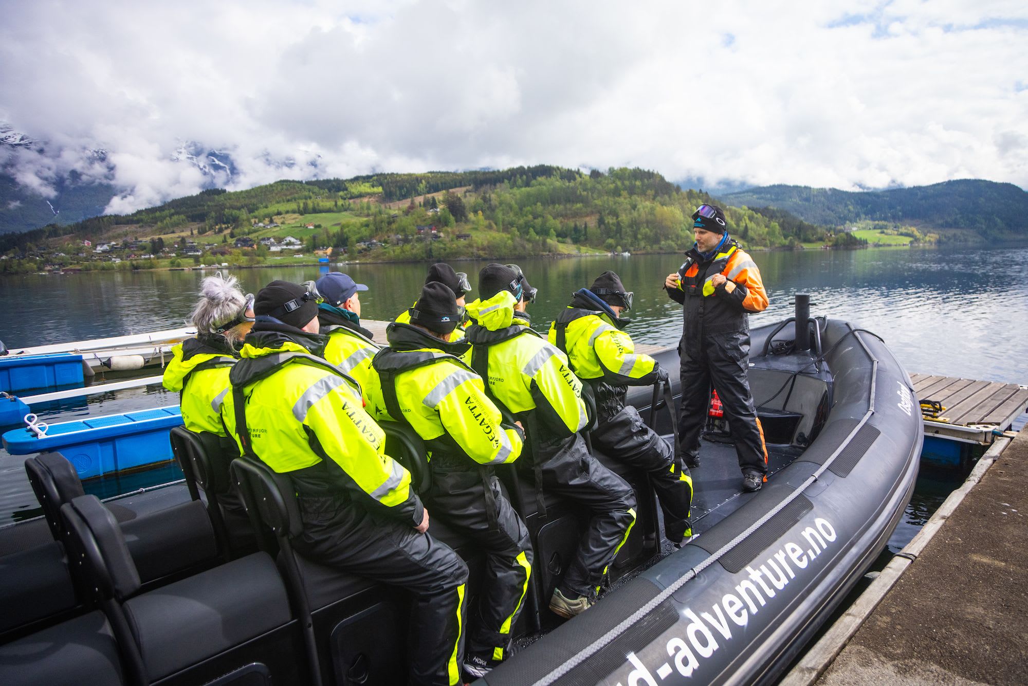 Guide in orange-black suit instructs tourists in yellow jackets aboard docked boat, scenic village behind.