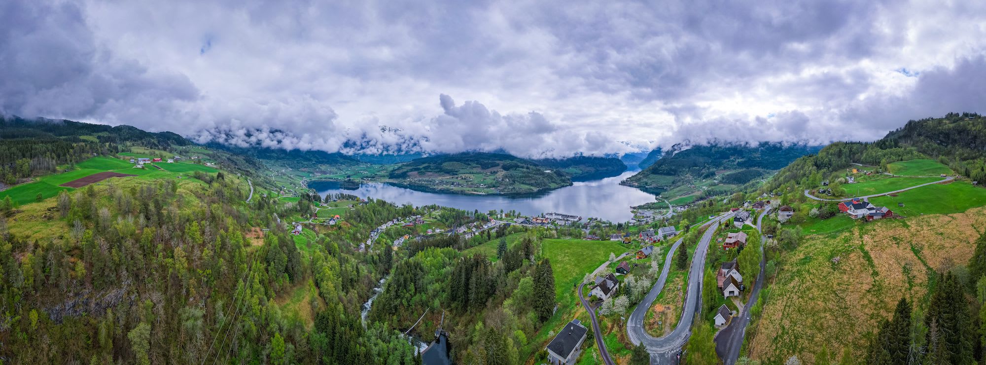 Panoramic aerial view of serene fjord landscape, lush farmland, charming houses, and misty mountains.