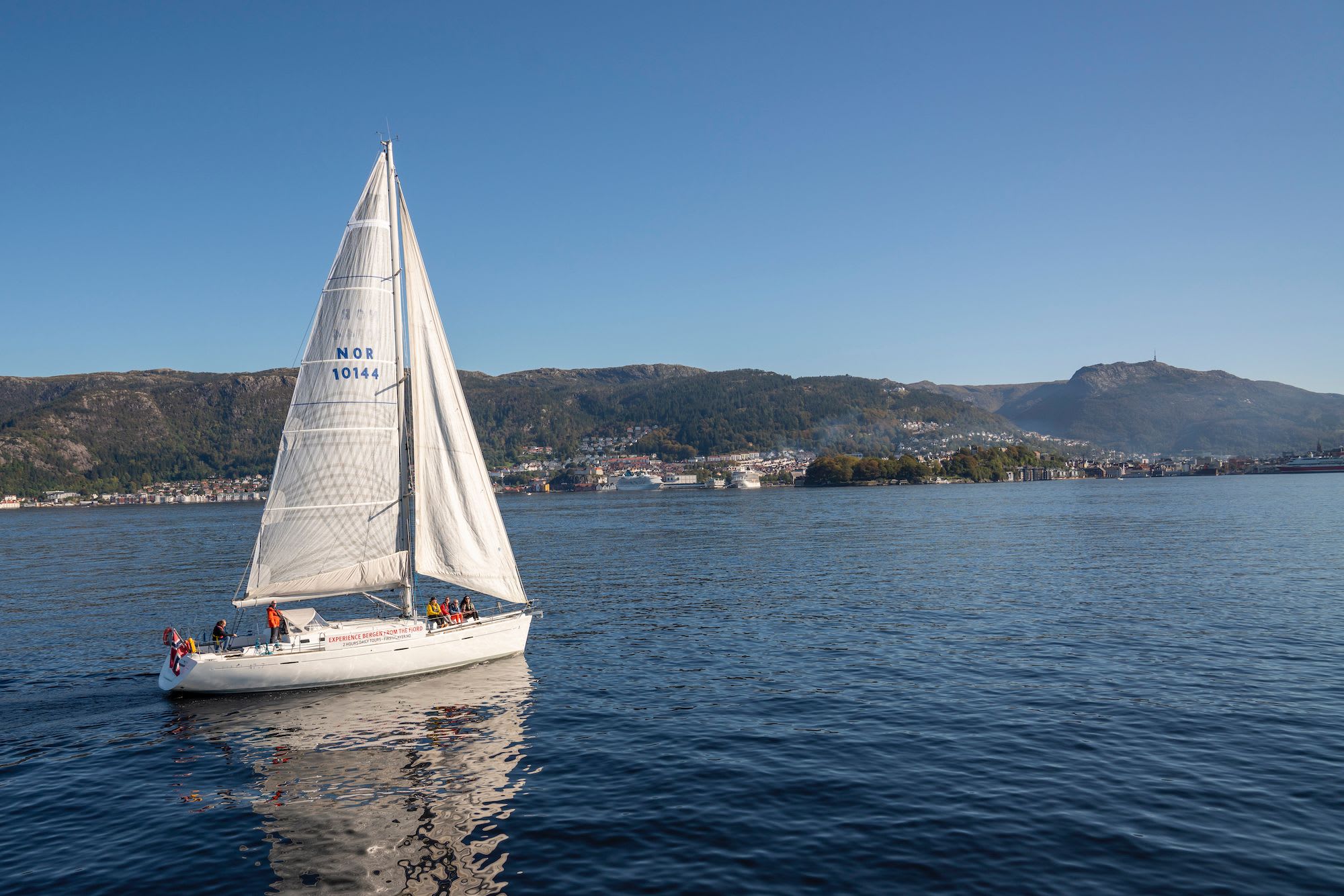 A sailboat with "NOR 10144" sails on calm waters with a mountainous landscape, clear blue sky, and distant coastal town.