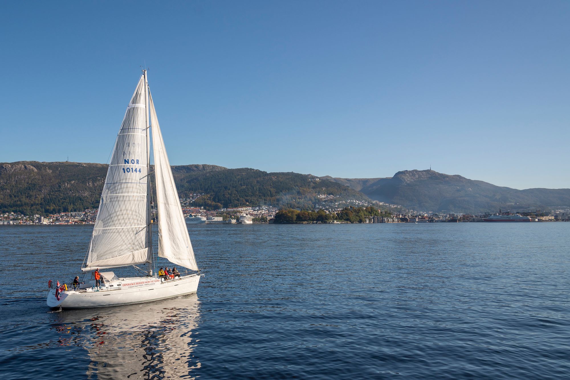 White sailboat with "NOR 1014-4" sail sailing under clear blue sky, people onboard, with Bergen city in the background.
