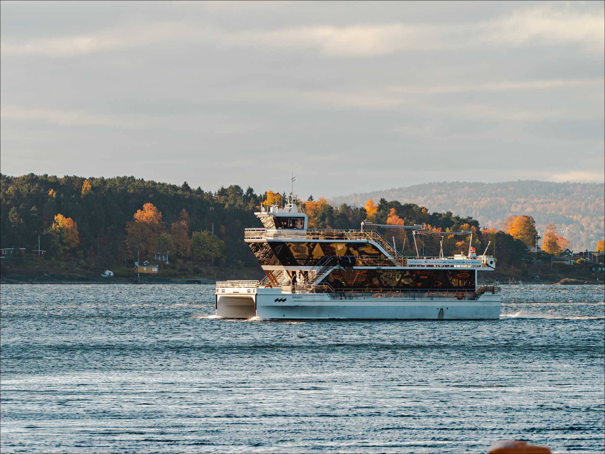 Hvit hybrid-elektrisk cruisebåt seiler gjennom Oslofjordens farvann med høstfargede skogåser