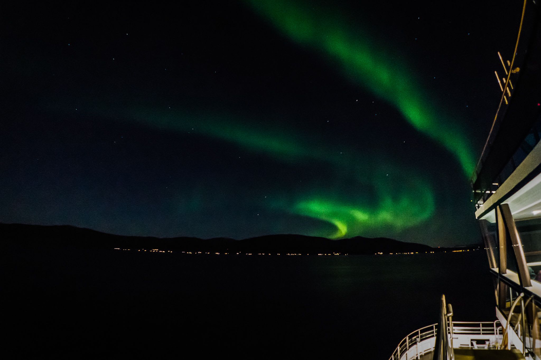 Et levende nordlys over fjellene, med grønne auroraer som virvler på himmelen, og byens lys til venstre.