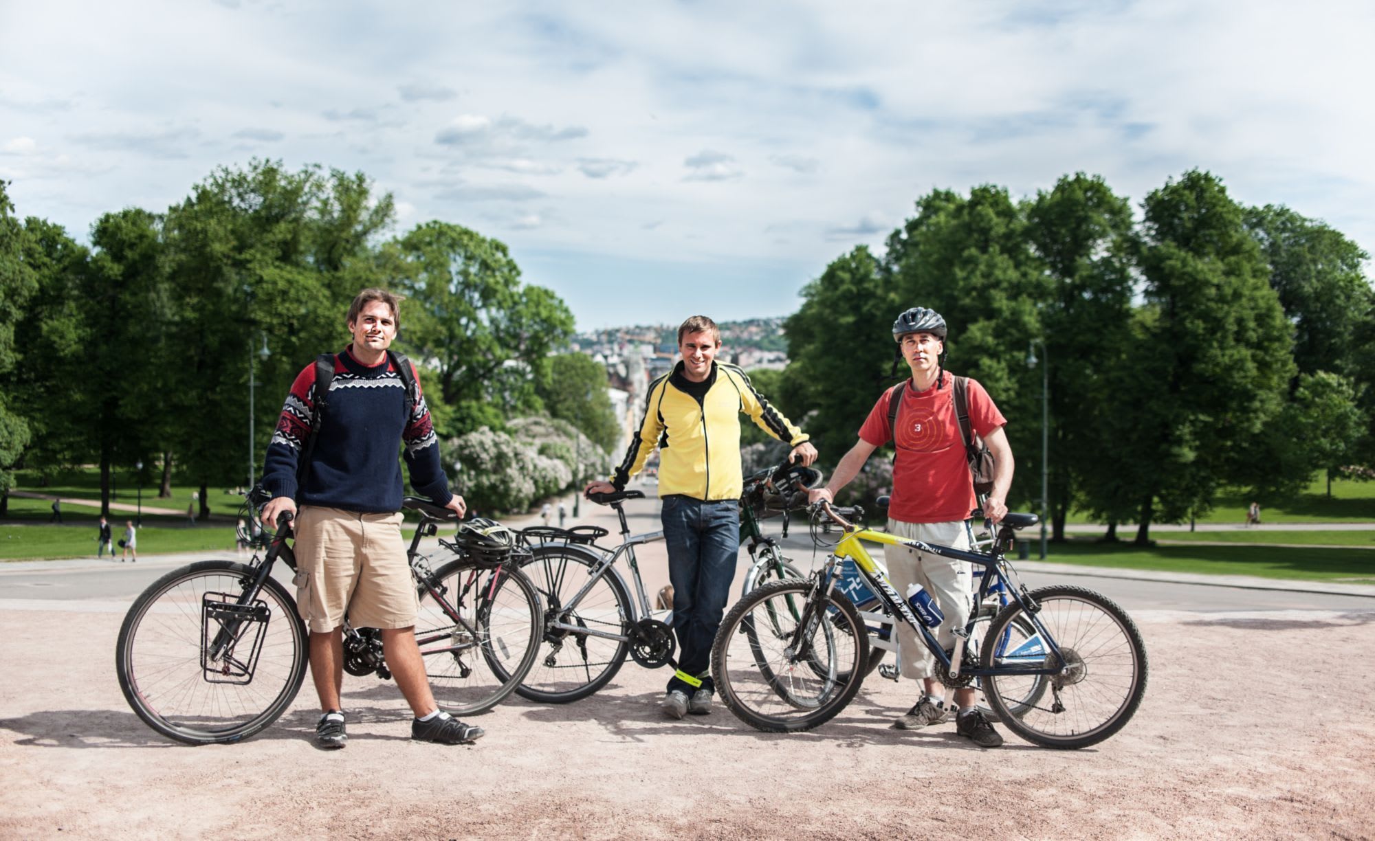 Tre menn poserer med sykler i park, byen i bakgrunnen.