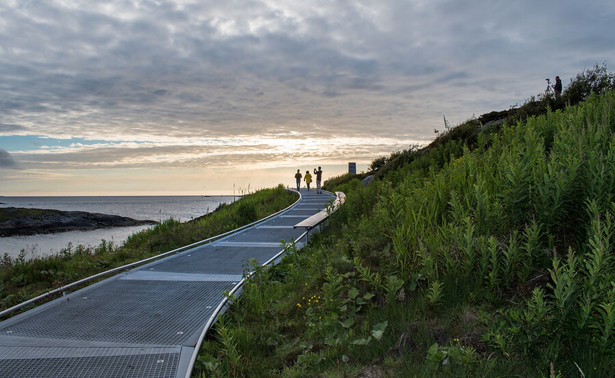 People walking along metal pathway towards horizon, with coastline and soft evening light.