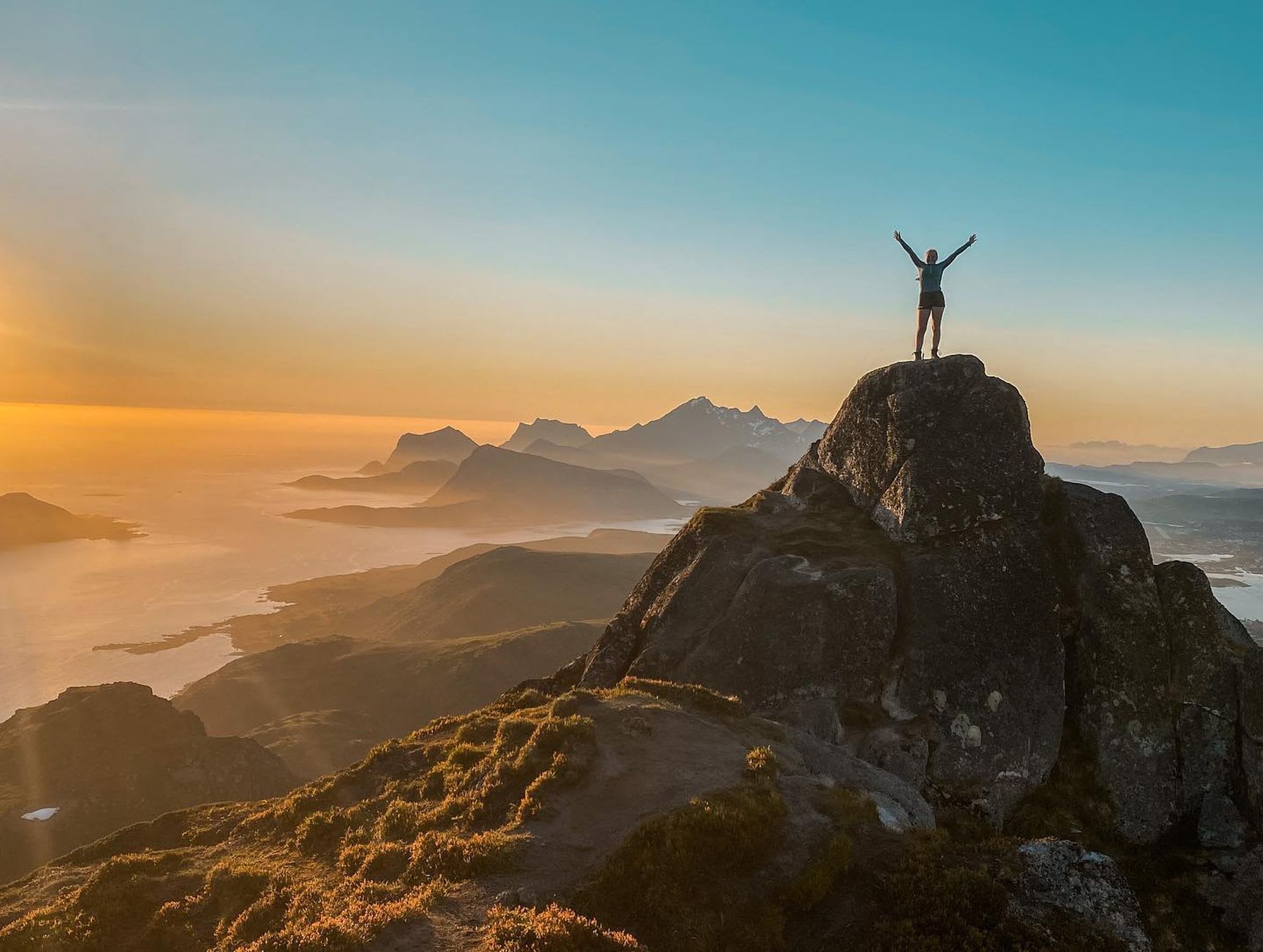 Person som feirer på toppen av en steinformasjon i Lofoten, med fantastisk kystlinje og solnedgangslys i bakgrunnen.