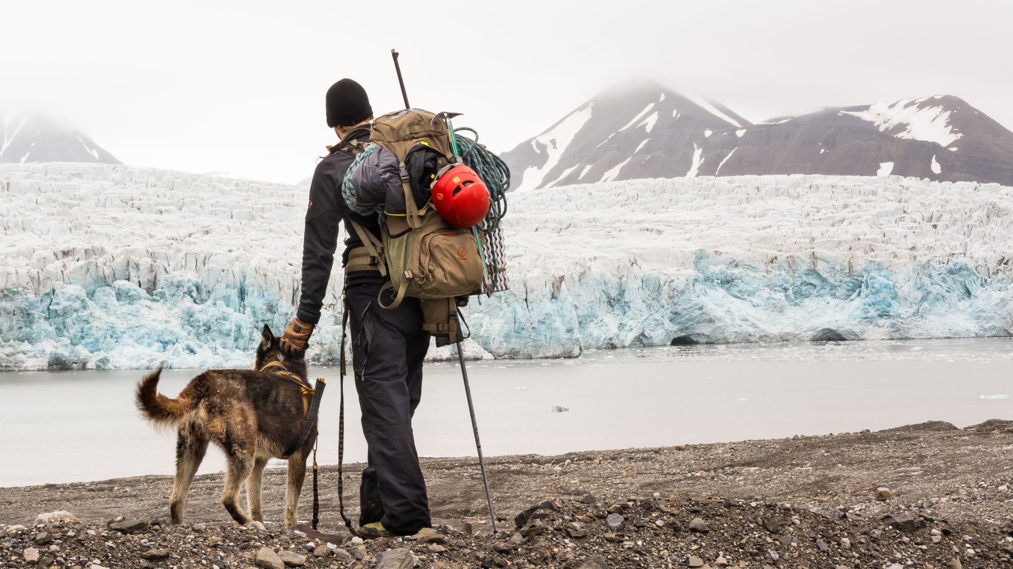 Climber on rocky shoreline with dog, gazing at glacier in the distance, preparing for ice climbing expedition.