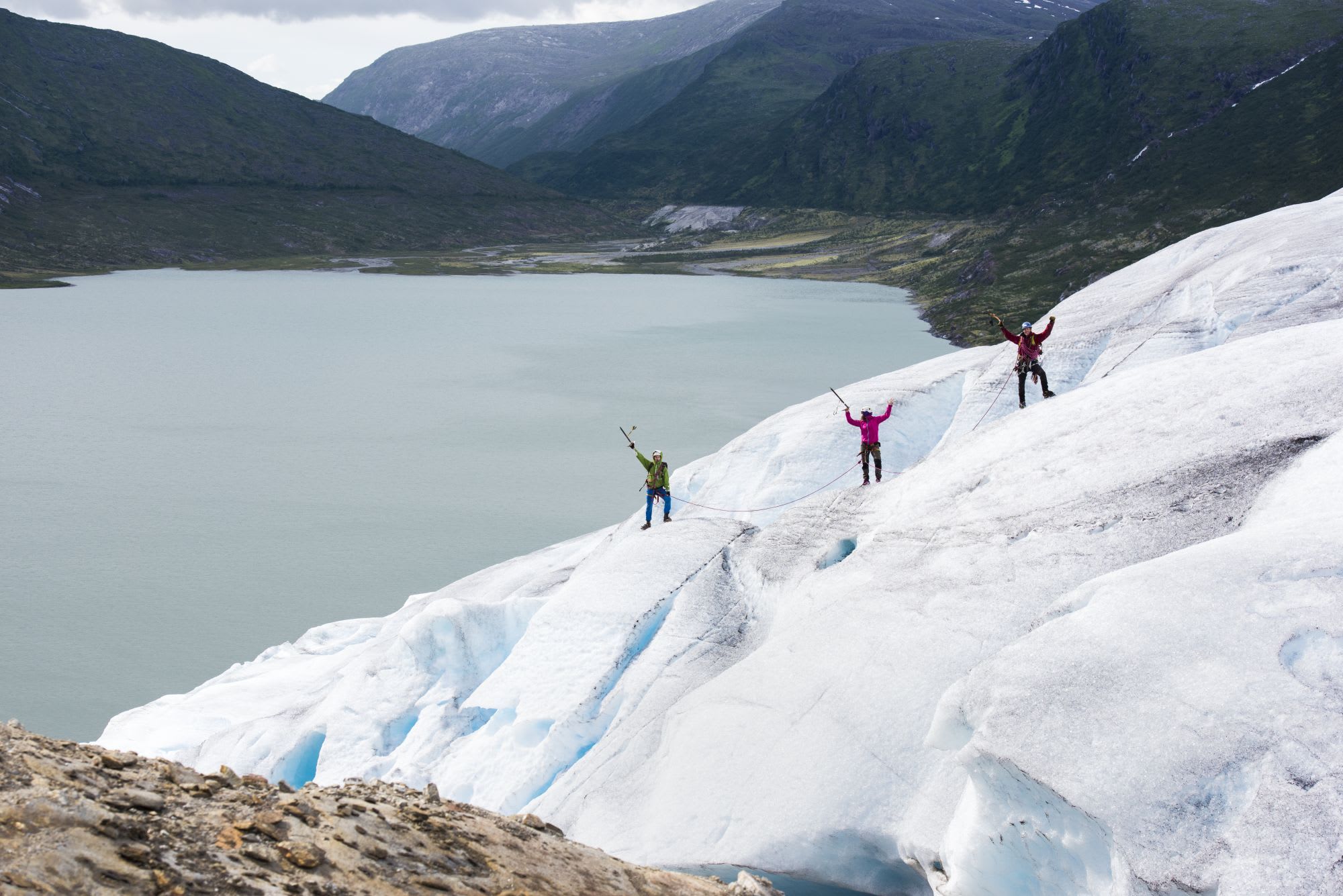 Three climbers on glacier, connected by rope, celebrating on icy surface with fjord and mountains in the background.
