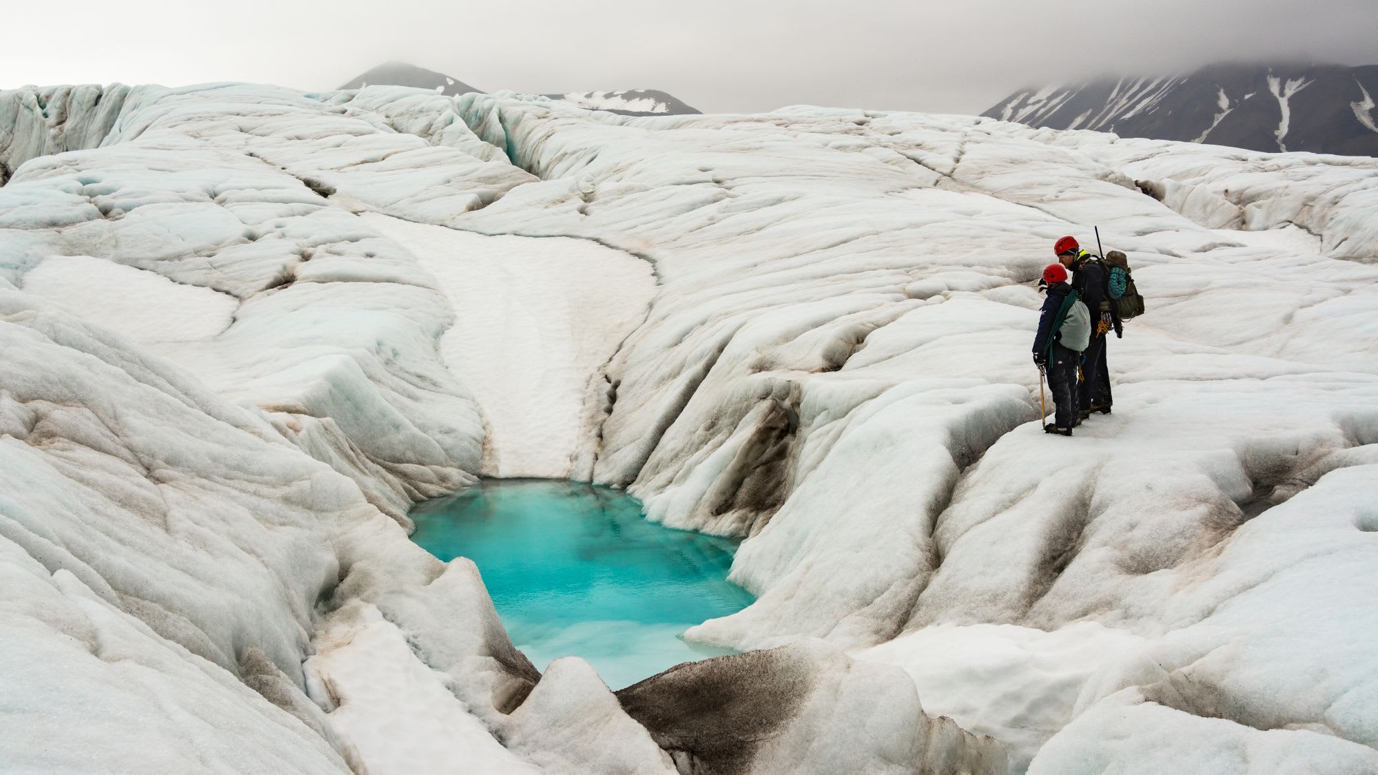 Two climbers standing on glacier surrounded by ice formations and crevasses, with turquoise pool and rugged terrain.