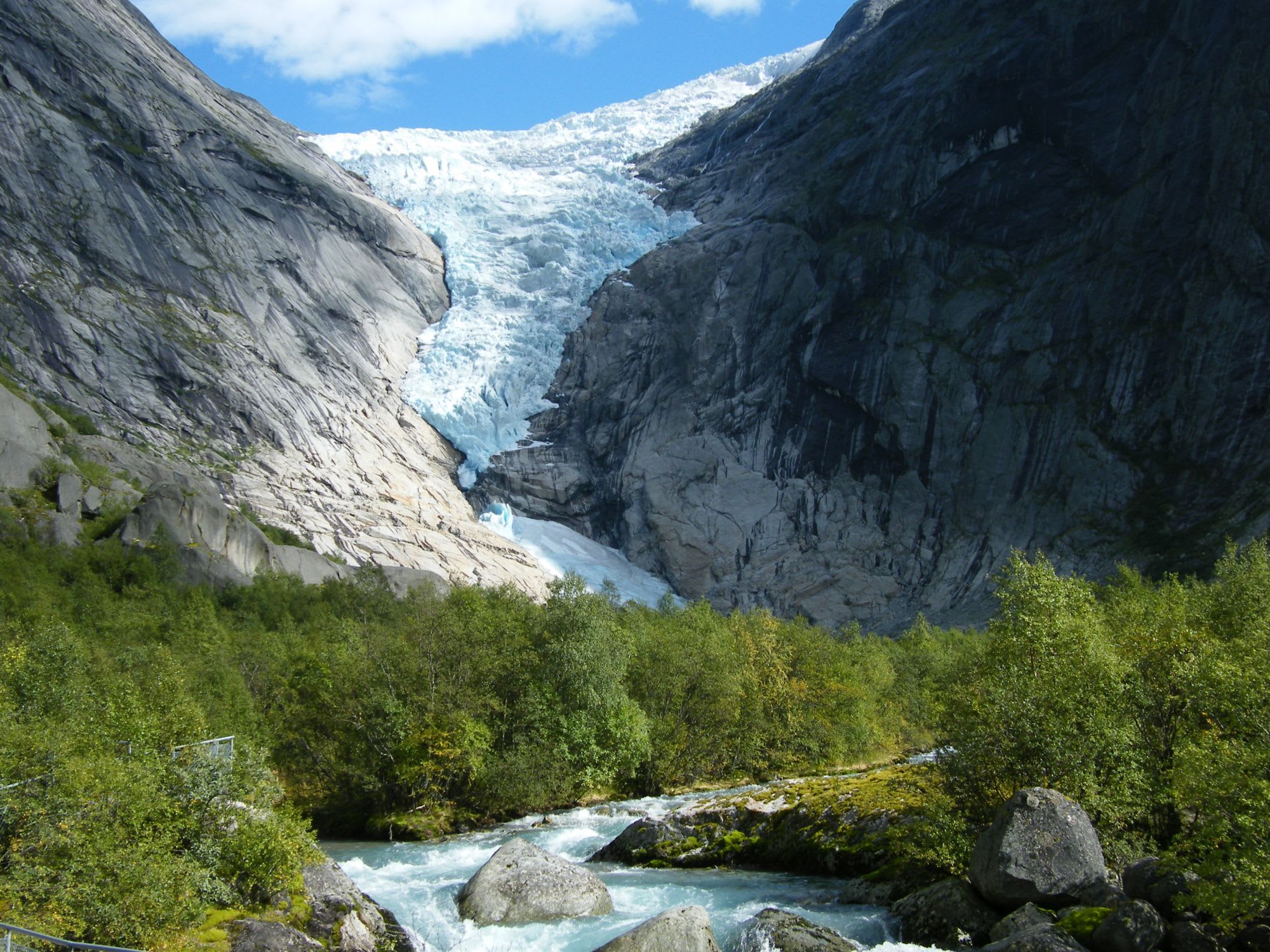 Glacier descending between towering cliffs, with blue ice, stream below, and lush greenery along the banks.