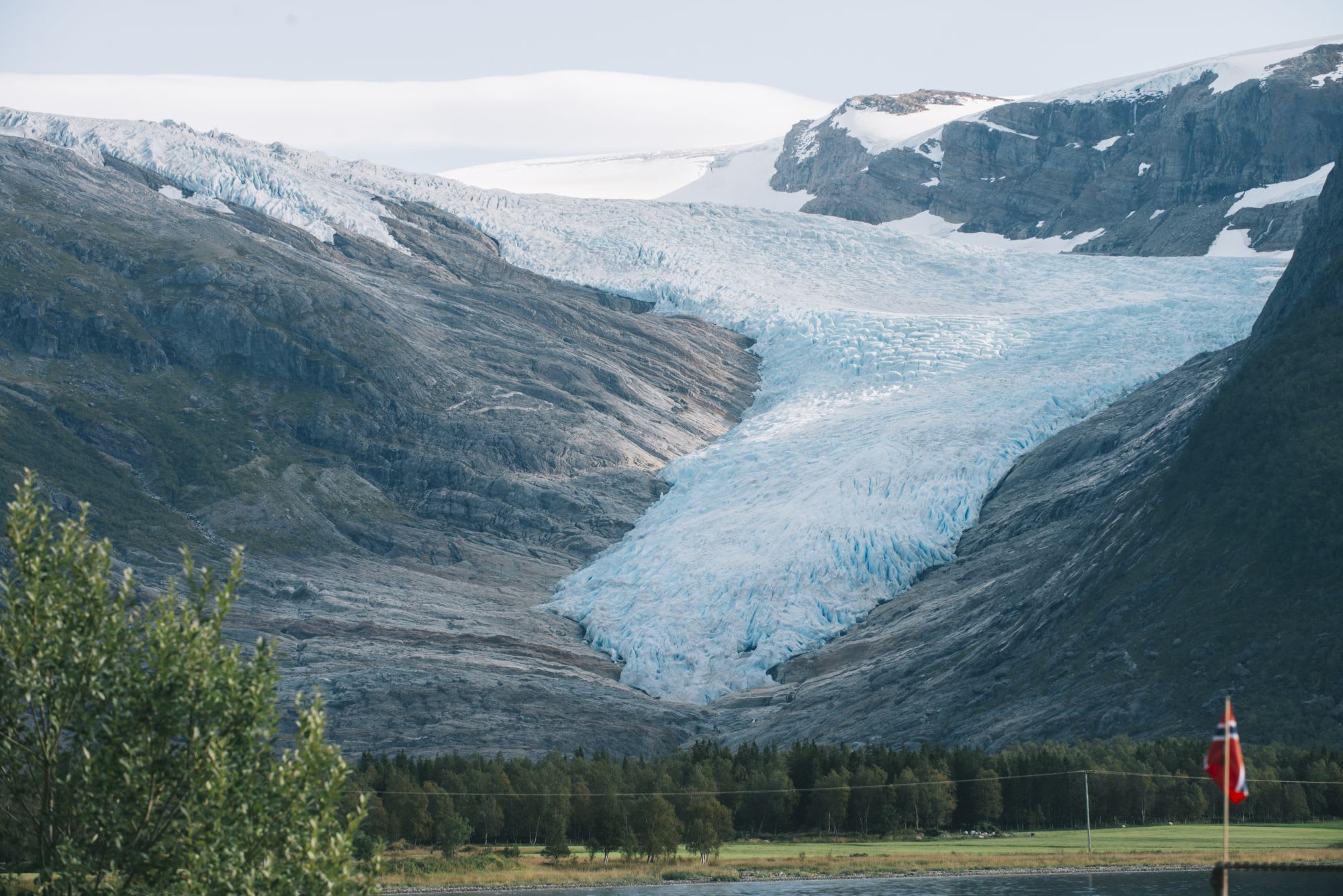 Stunning view of glacier flowing down from mountains with flag in foreground, green trees and snow-capped peaks.