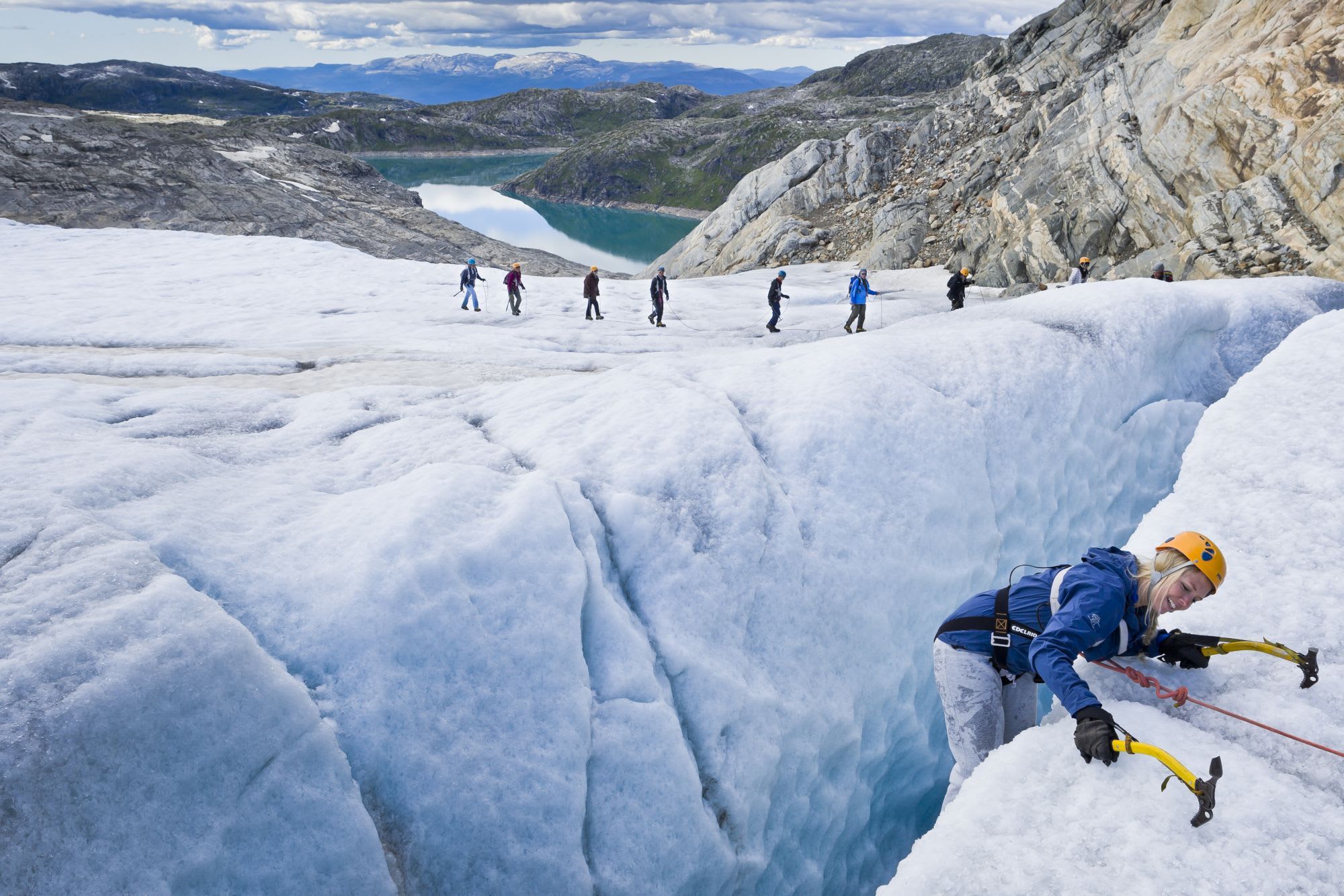 En kvinne klatrer ut av en sprekk og smiler, mens ni fjellklatrere går over en isbre med fjorder og fjell i bakgrunnen.
