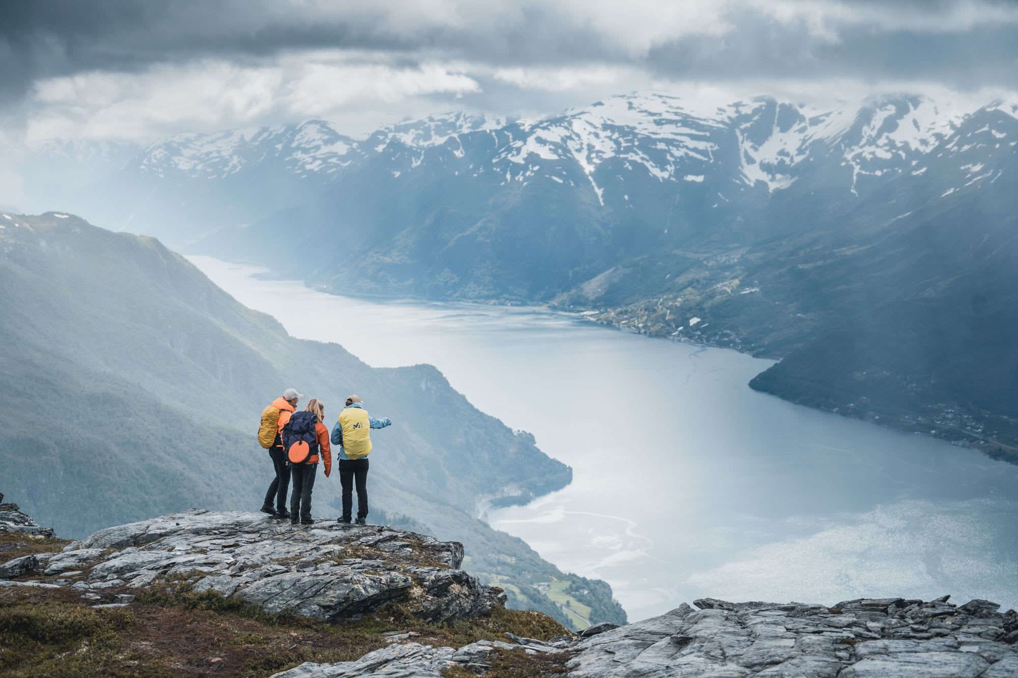 Three hikers on rocky outcrop overlooking misty fjord with snow-capped mountains, cloudy sky, and soft light.