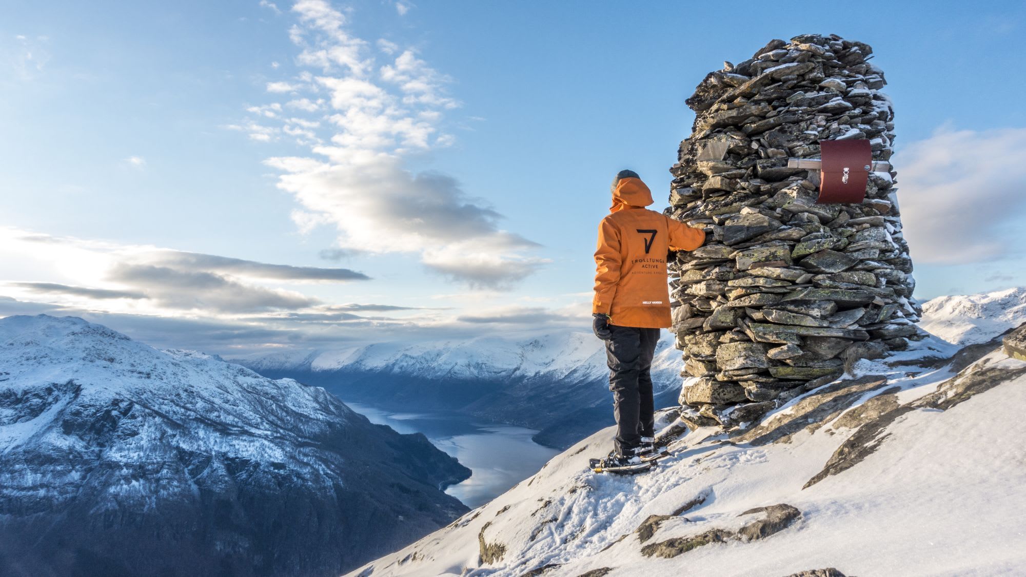 Turgåer i oransje jakke og snesko ved steinbunke som ser på snødekte fjell og fjord under klar himmel.