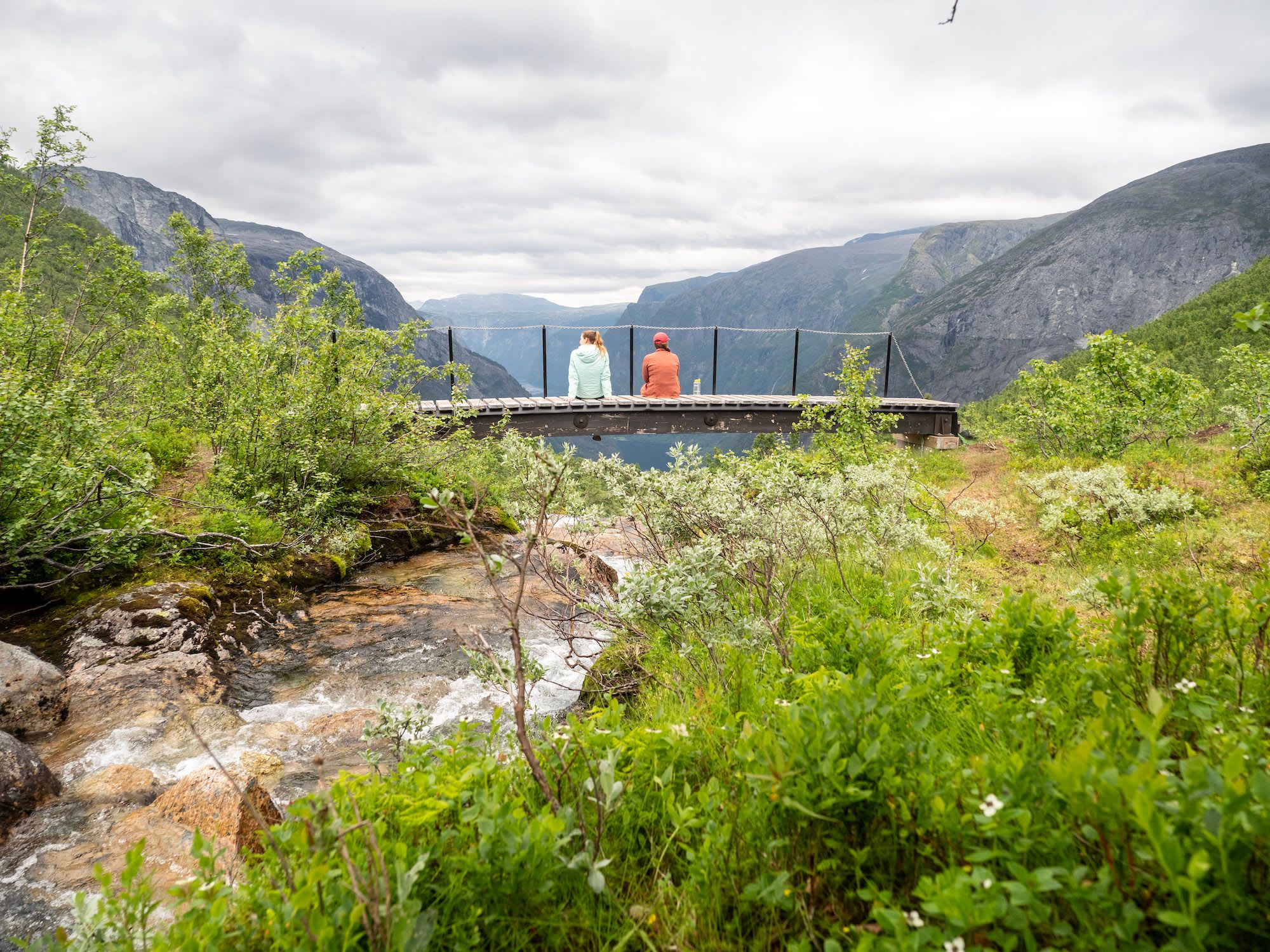 To personer som sitter på utsiktsplattform og ser ut over dalen med bratte, steinete fjell og rennende bekk under skyet himmel.