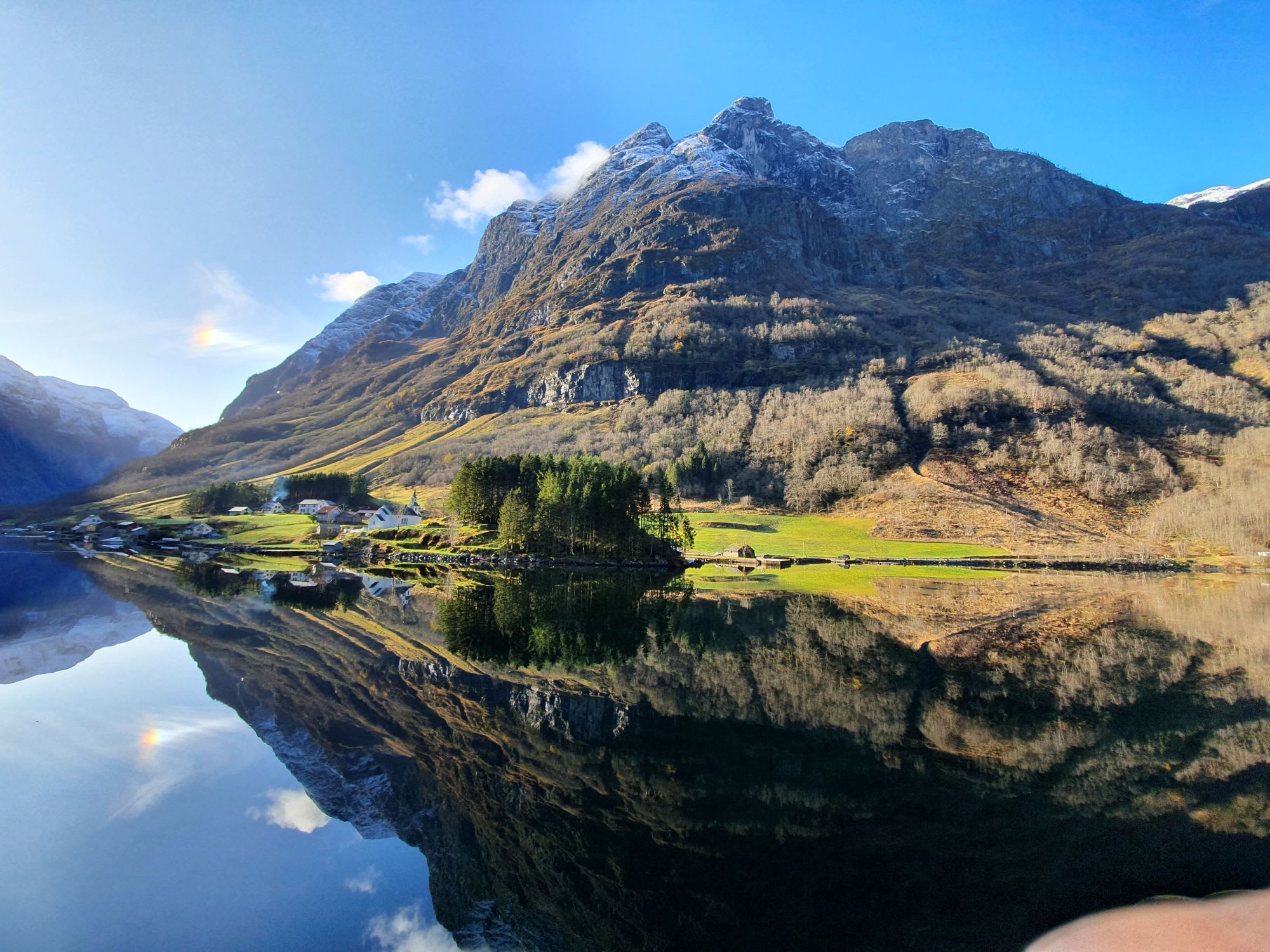 Rolig fjord med speilaktig vann som reflekterer steile fjell, snø og frodige hus langs kysten.