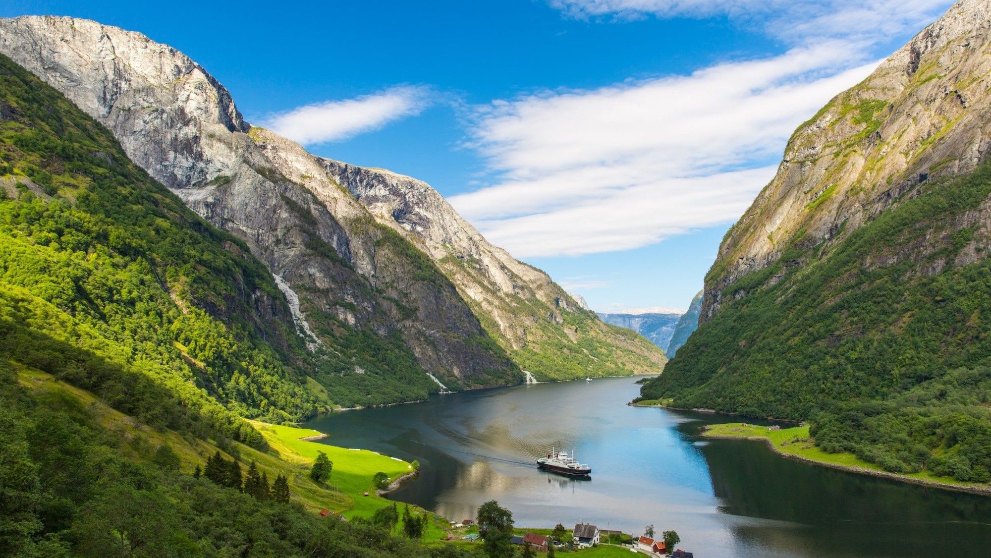 Fjord omgitt av steile klipper med livlige grønne marker og røde hustak under klar blå himmel.