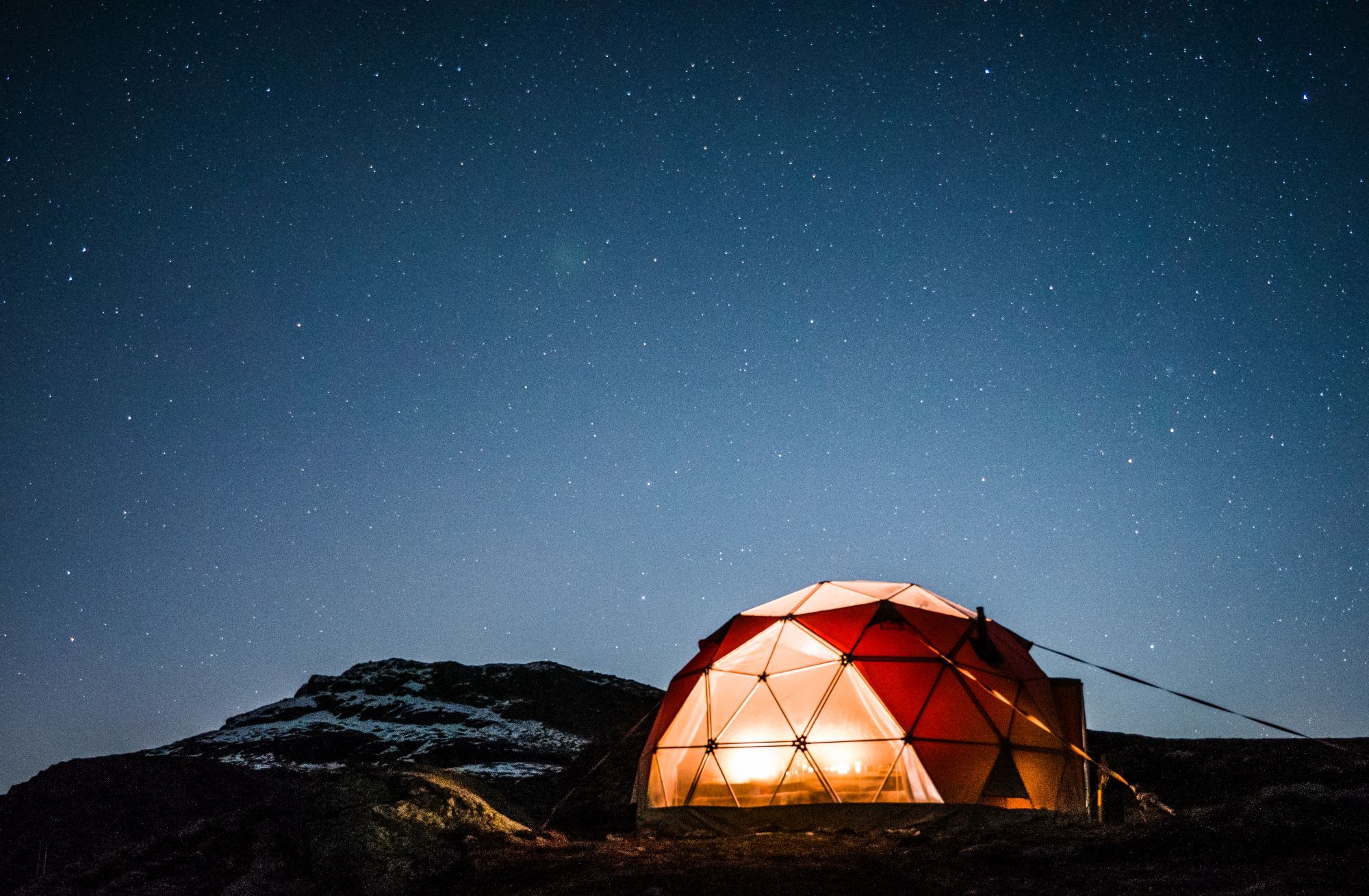 Geodesic dome tent illuminated from within, set against starry night sky with snow-capped mountains.