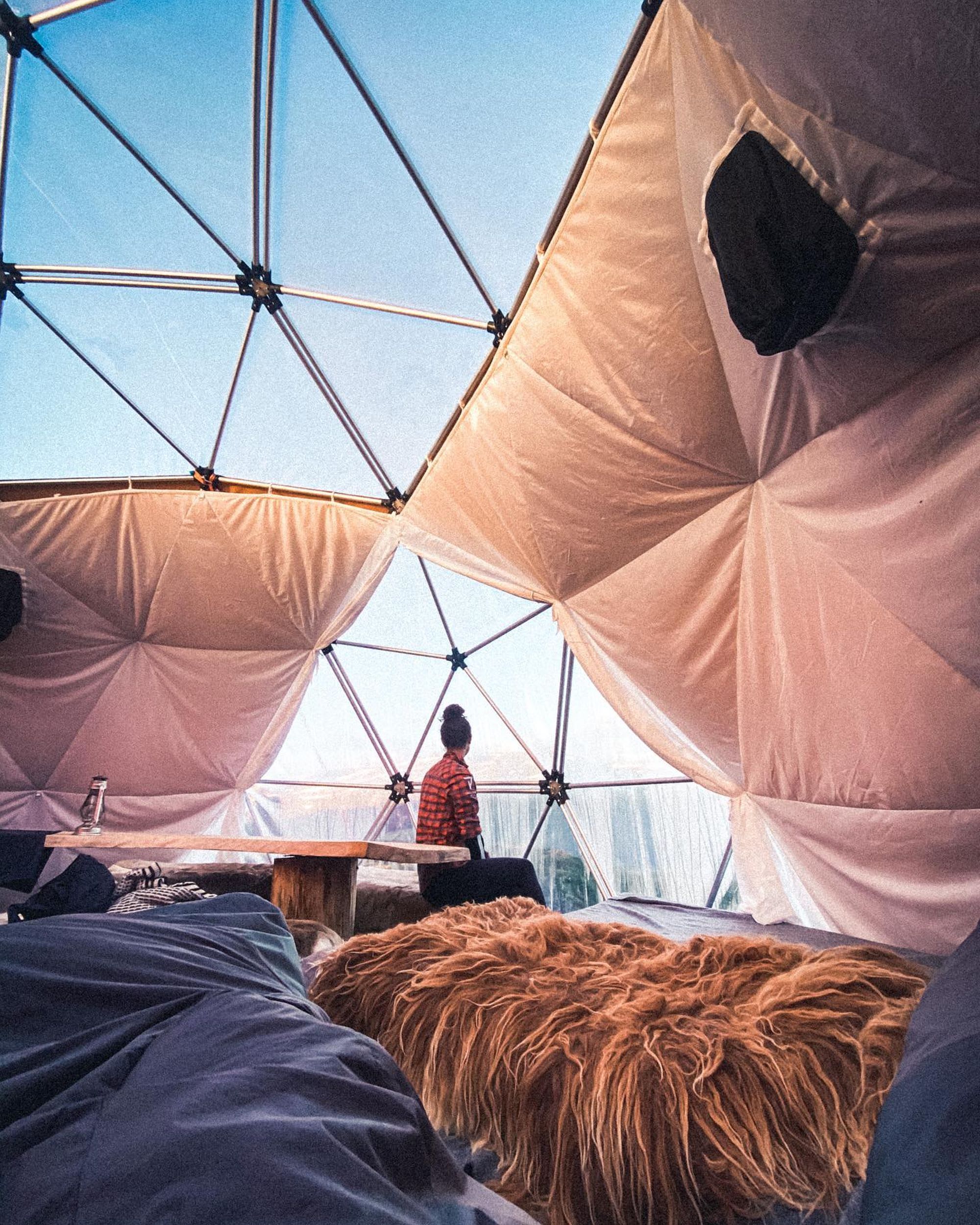 Person som sitter i koselig geodetisk kuppeltelt og ser ut av vinduet på naturskjønn utsikt med pelspledd og trebord.