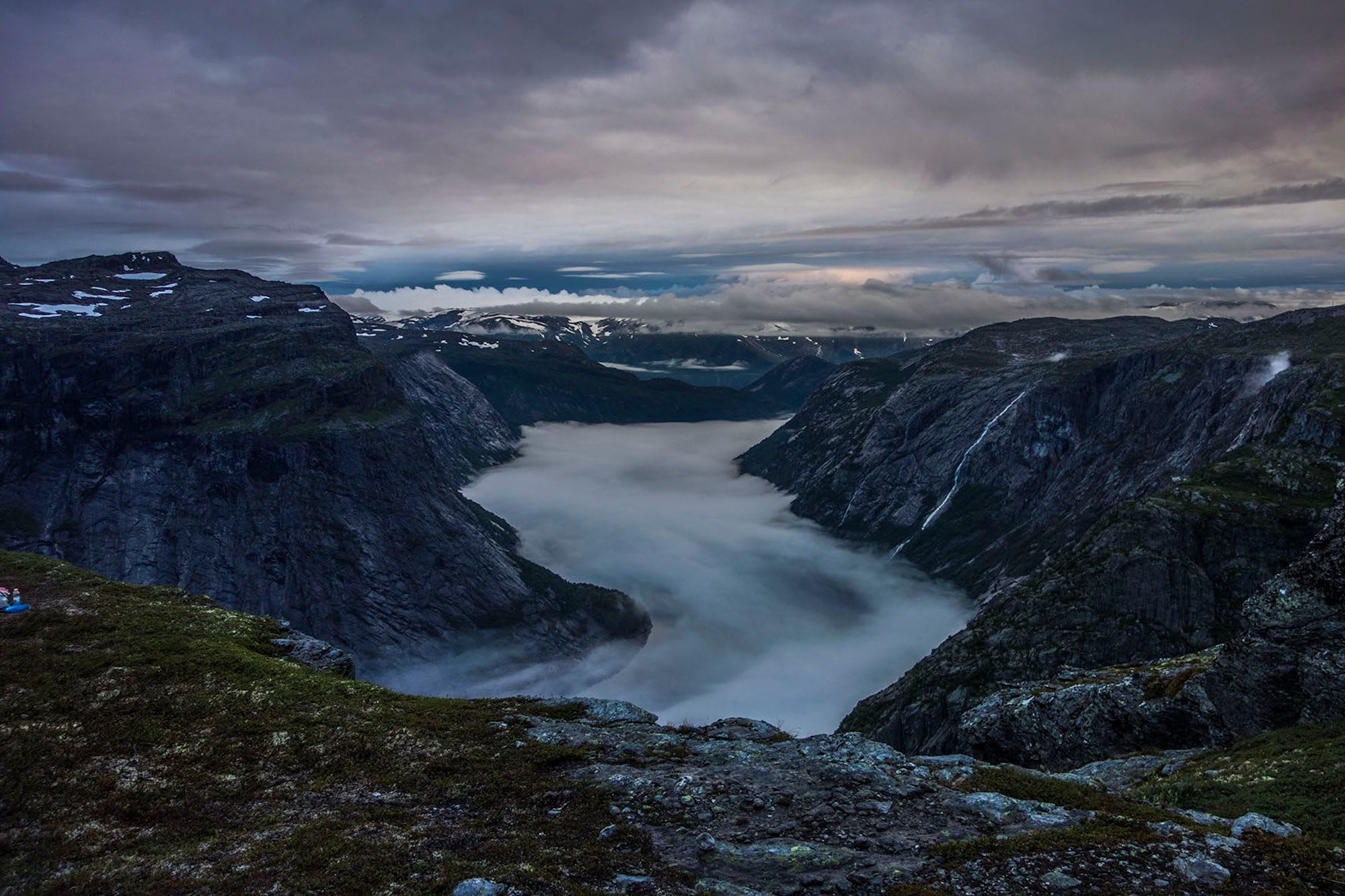 Misty fjord with fog covering the water, snow-capped mountains, and grey clouds above.