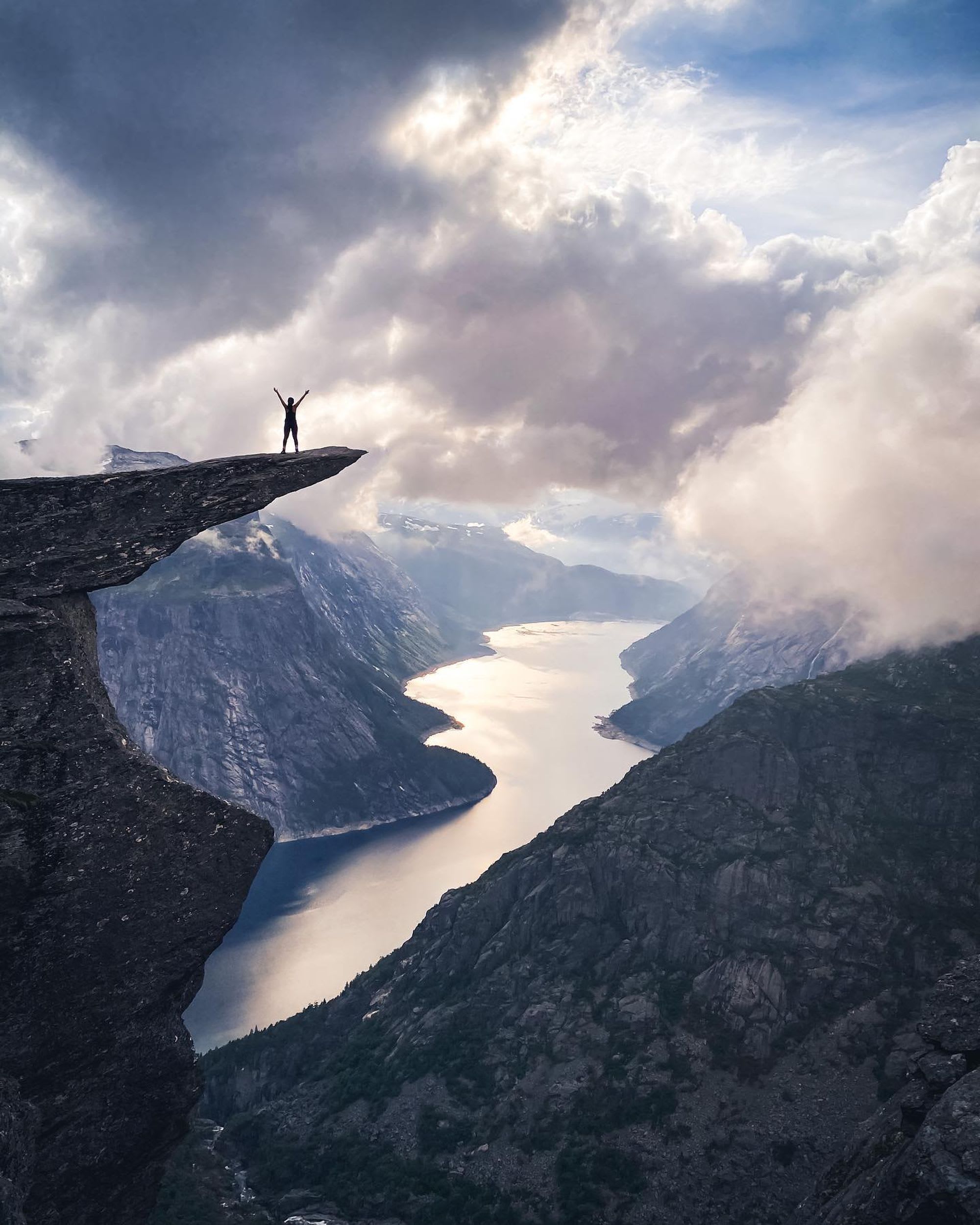 Person med armene hevet som står på klippekanter, silhuett mot dramatisk himmel og fjord omgitt av fjell.