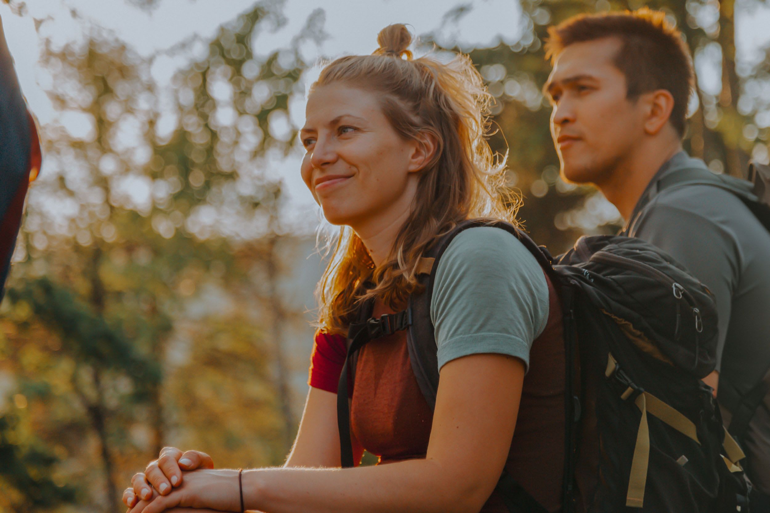 To smilende turgåere hviler i skogen i gyllen time, sollys filtrerer gjennom grønne trær.