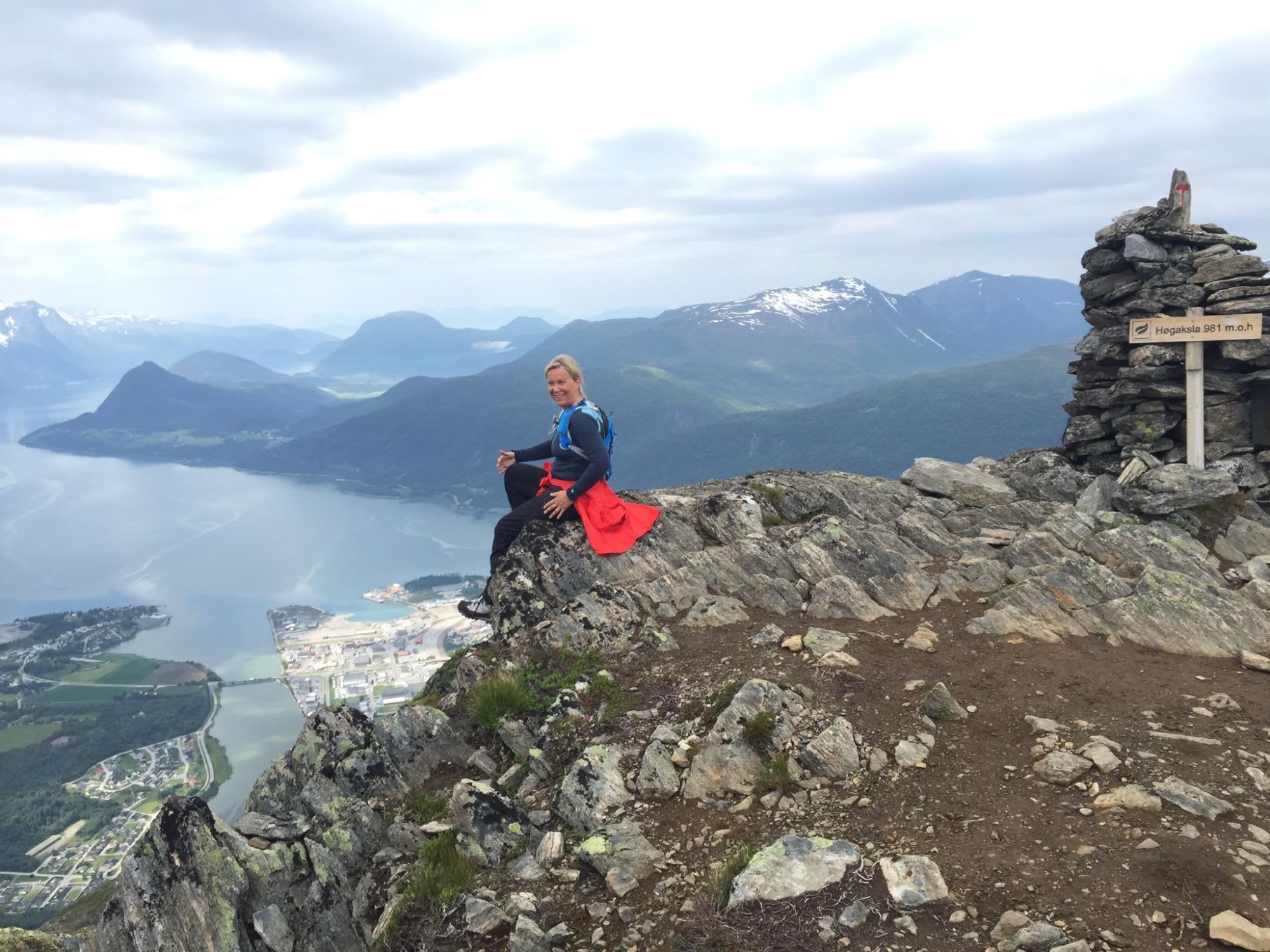 Smilende kvinne sitter på en klippe ved skiltet "Høgaksla 981 m.o.h.", byen nedenfor.