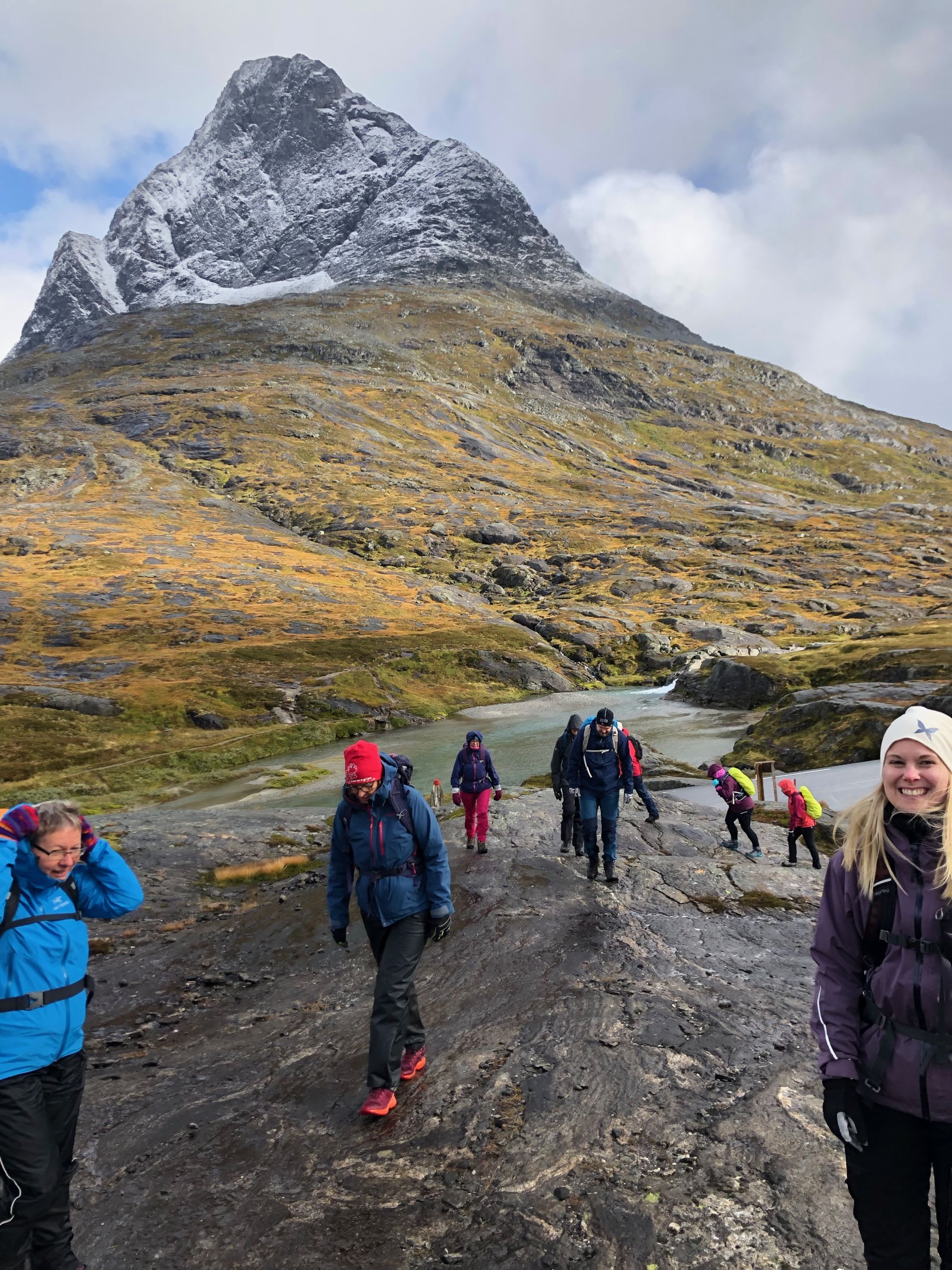 Hikers walking across rocky terrain with snow-capped mountain in the background, some resting and others trekking.
