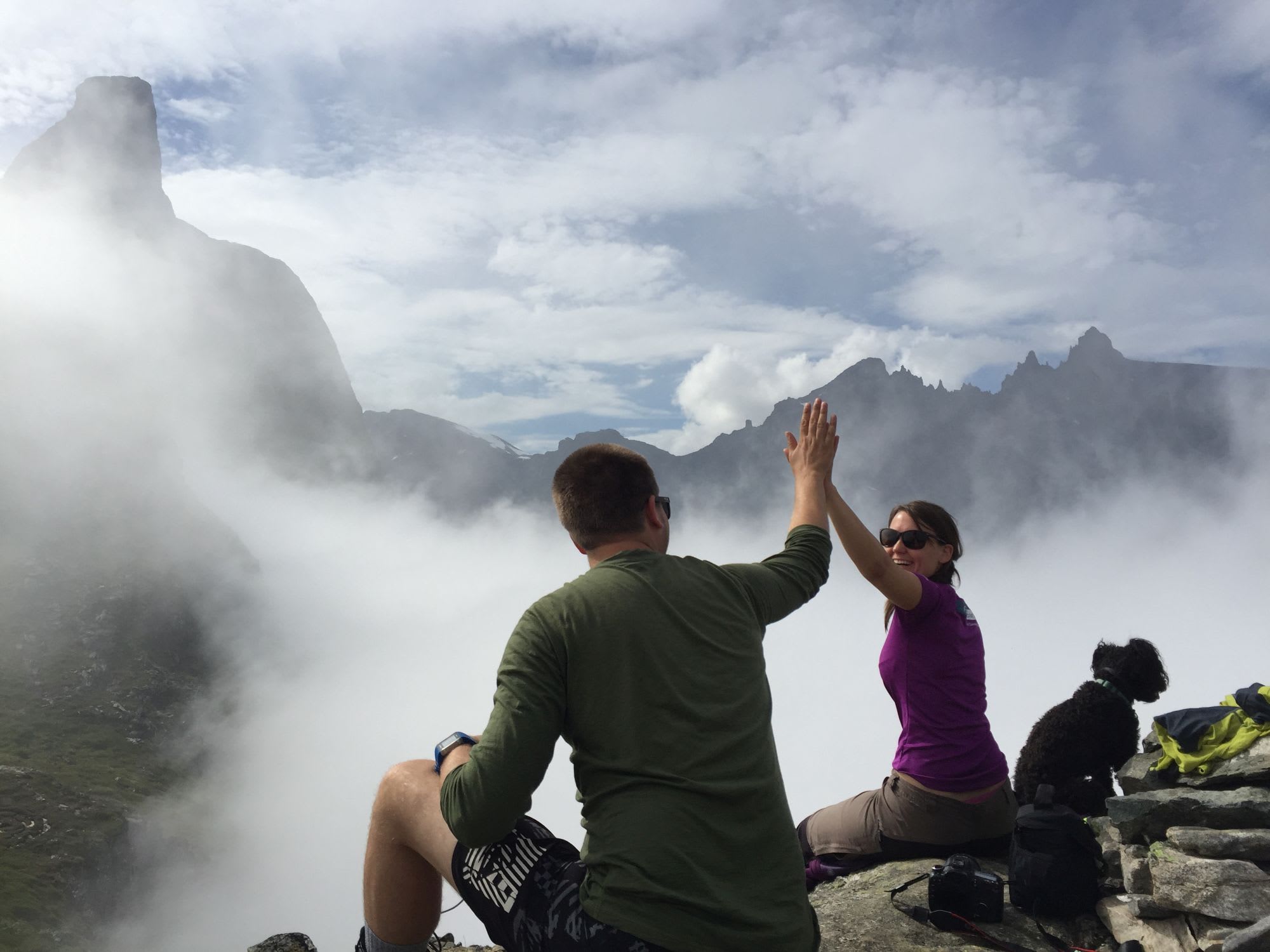 Two hikers sitting on rocky ledge above clouds, giving each other joyful high-five with a dog nearby.