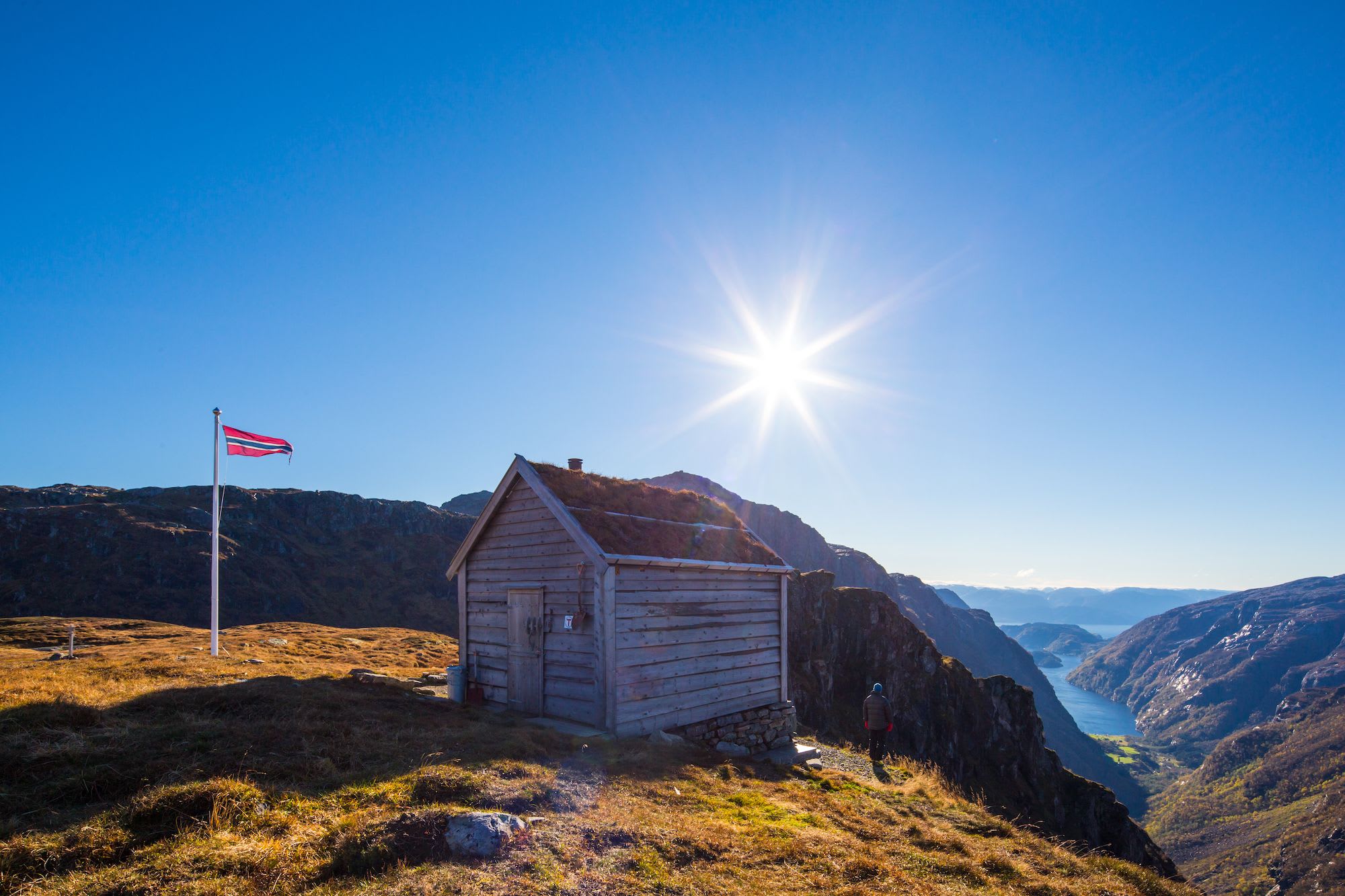 Small wooden cabin at Kiellandsbu with grass roof on sunlit hillside, overlooking rugged valley and calm fjord