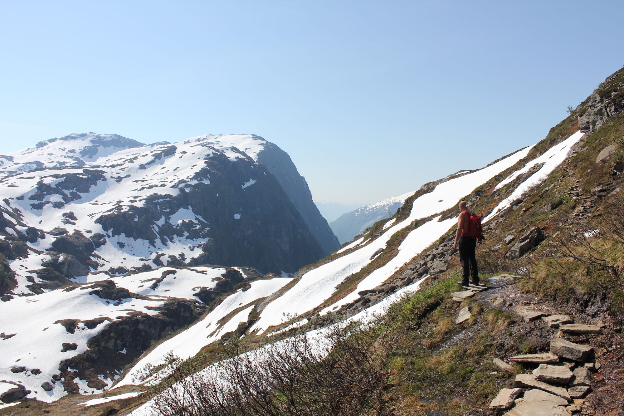 Hiker with red backpack on rocky mountain trail, overlooking deep valleys and snow-covered peaks.