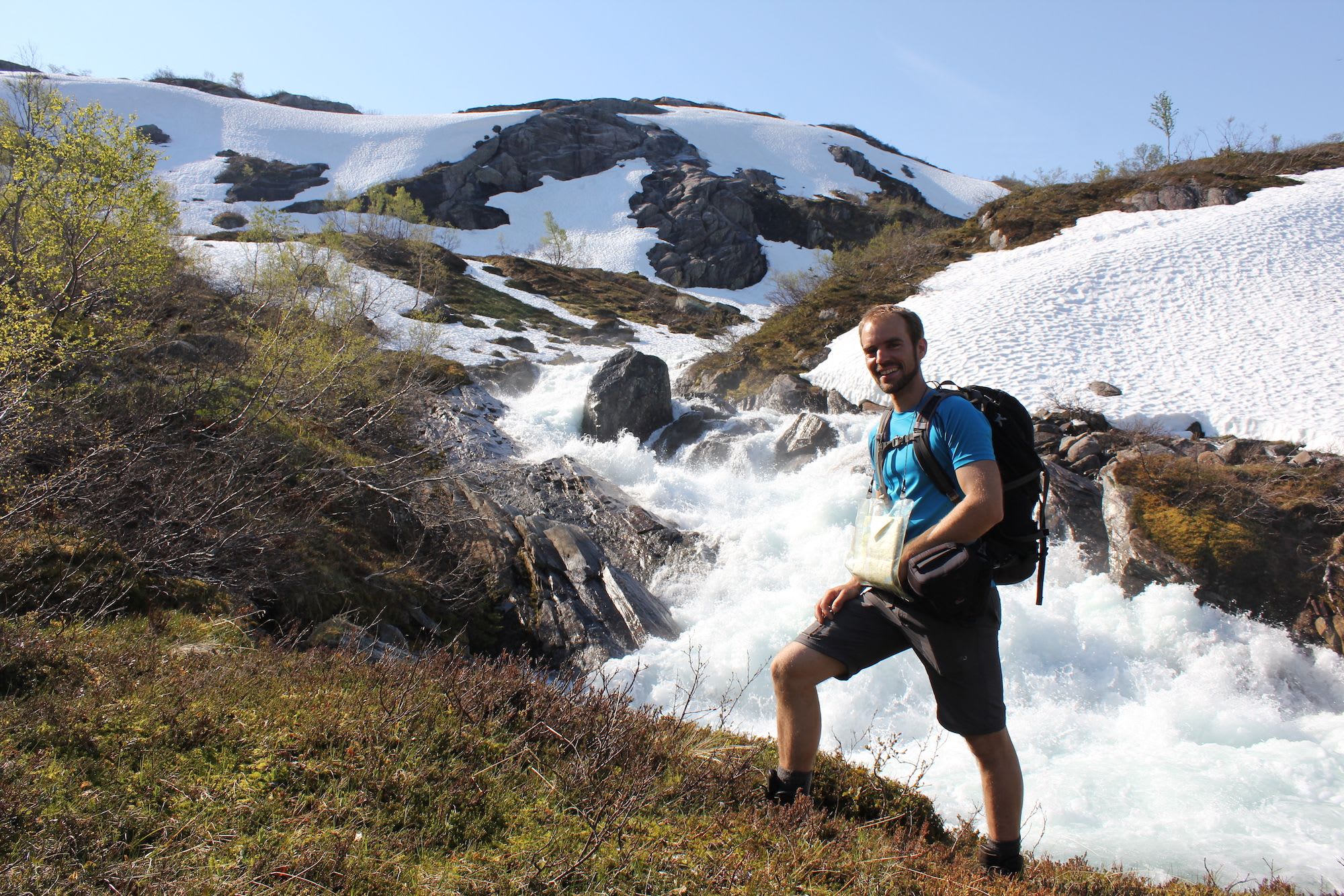 Smiling hiker in blue shirt and backpack stands by foamy mountain stream, with rugged landscape and melting snow.