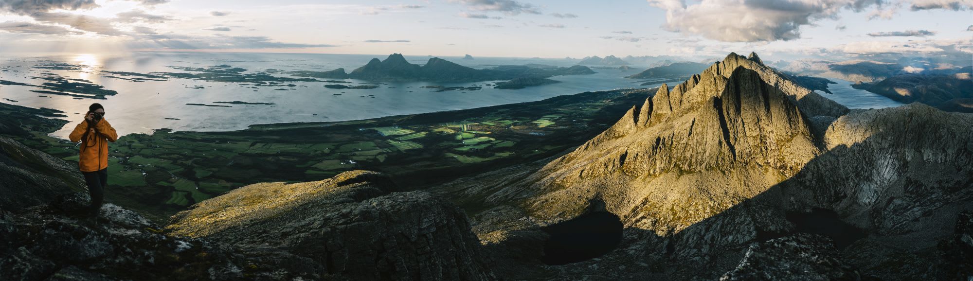 Photographer in yellow jacket capturing coastal landscape with dramatic peaks, green farmlands, and calm sea.