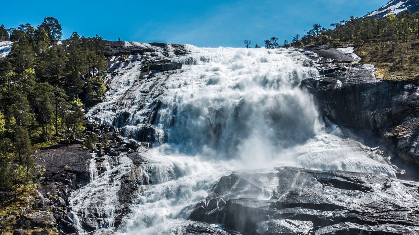 Solbelyst foss som faller over steinete terreng med omkringliggende grøntområder.