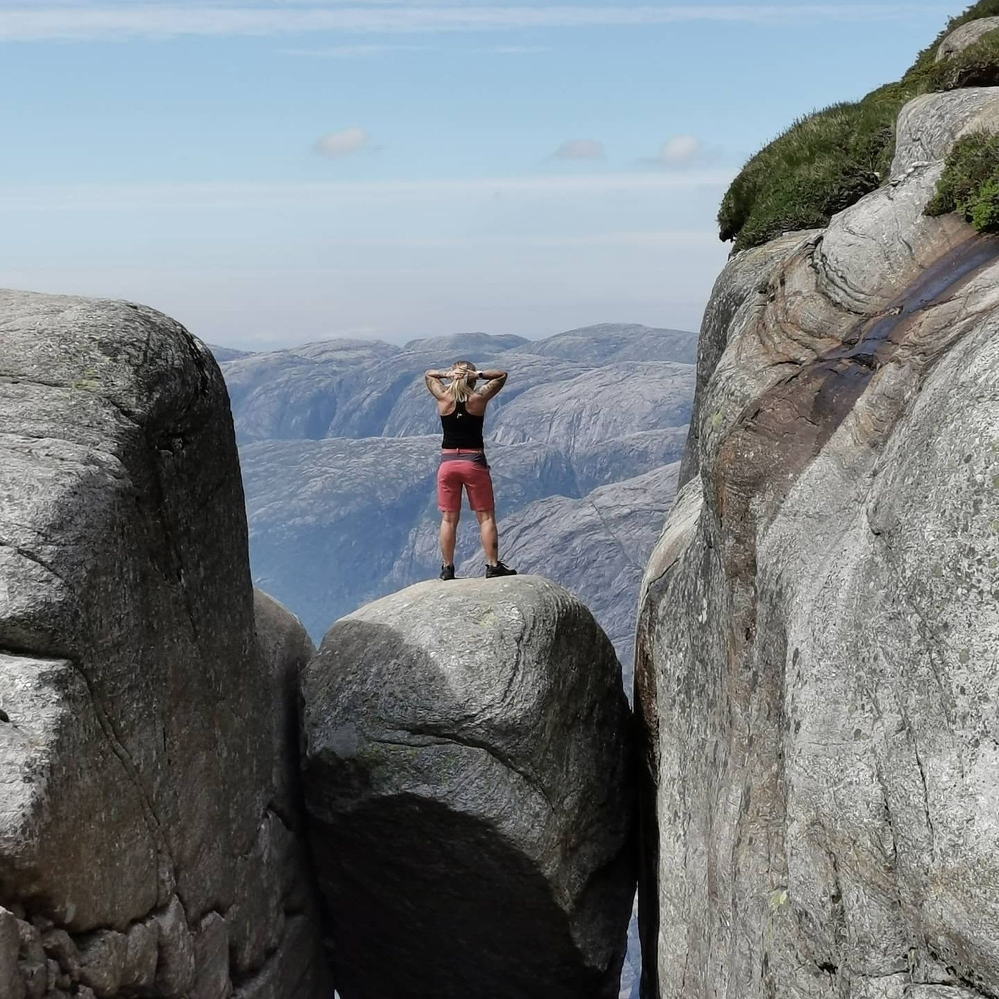 Woman standing on Kjeragbolten, gazing at breathtaking landscape with hands on her head, surrounded by cliffs.