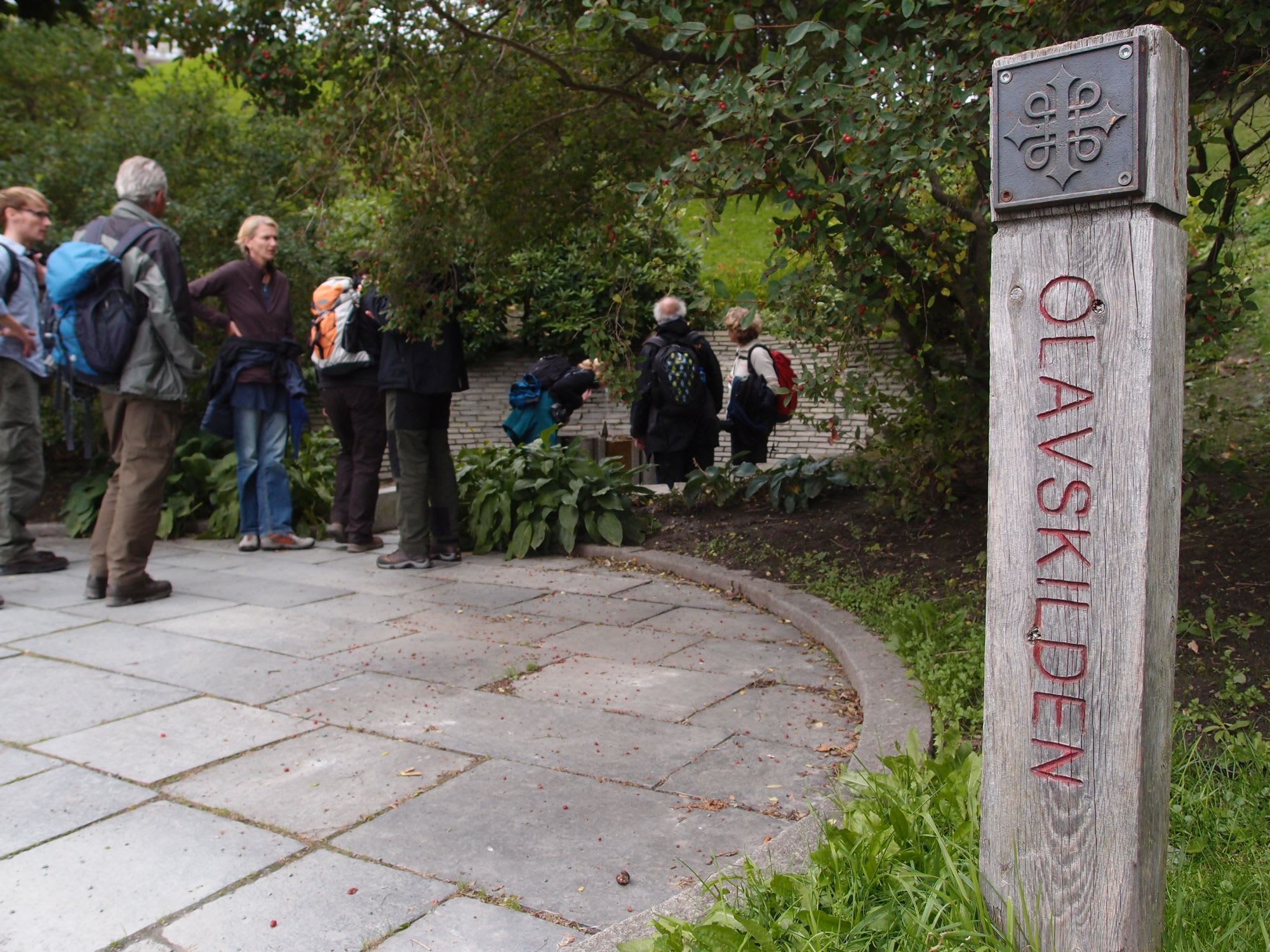 Gruppe turgåere nær “Olavskilden” kilde, med et trepost med korssymbol, omringet av grønn vegetasjon og steinsatte stier.