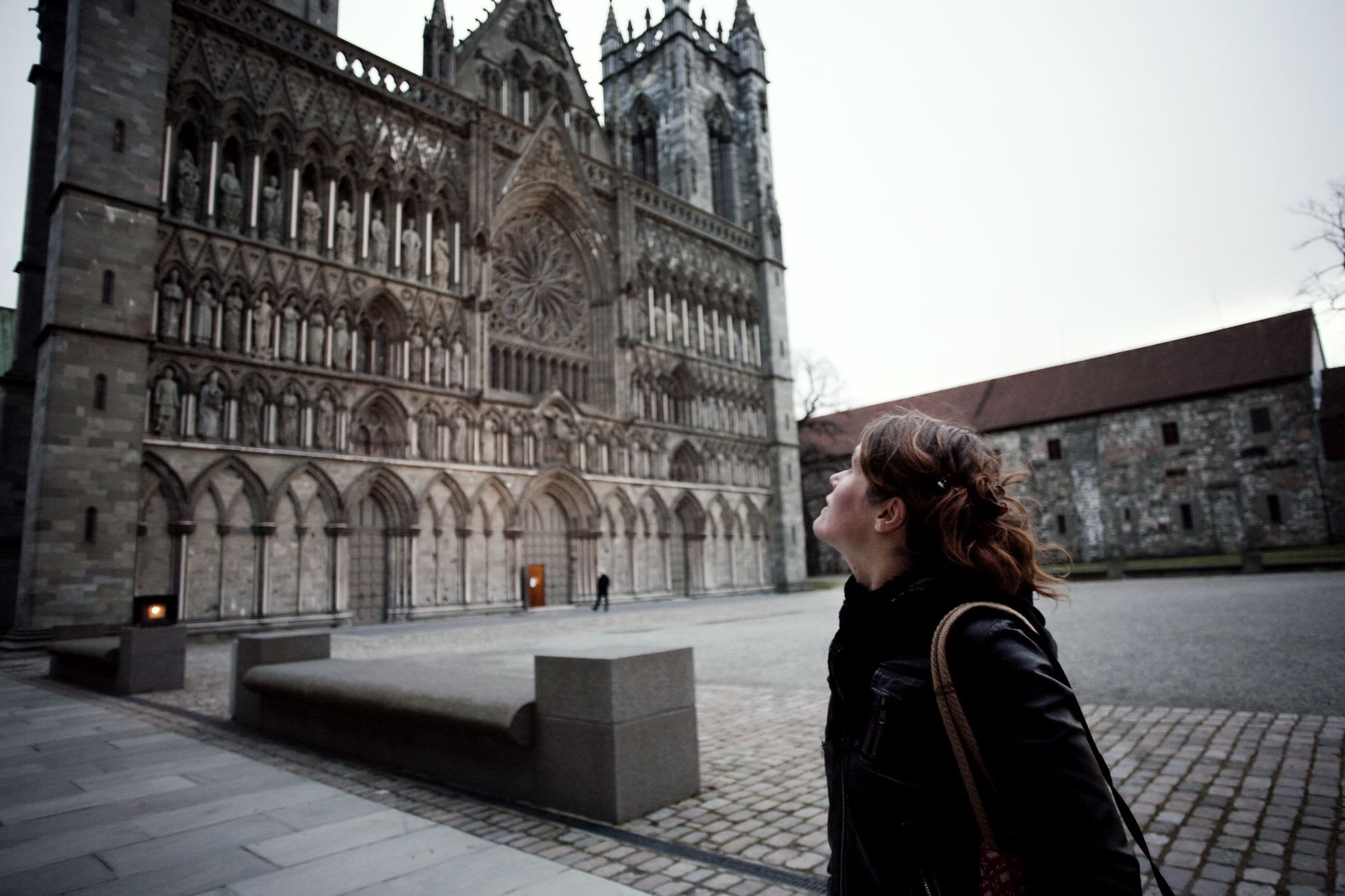 Kvinne som ser opp på den intrikate fasaden til Nidarosdomen med detaljerte utskjæringer og statuer, under et overskyet himmel.