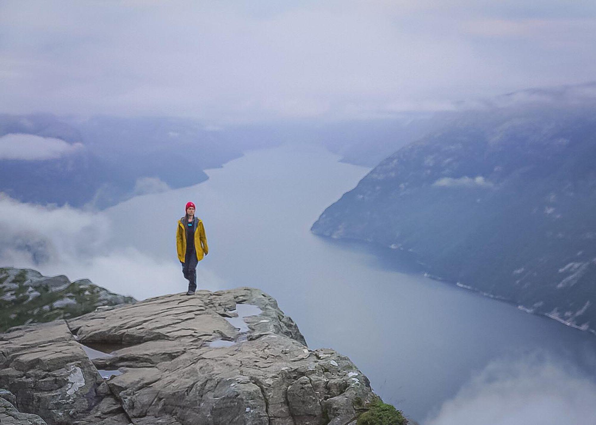En person i gul jakke og rød lue står på en klippe og ser ut over en rolig fjord og tåkete fjell.