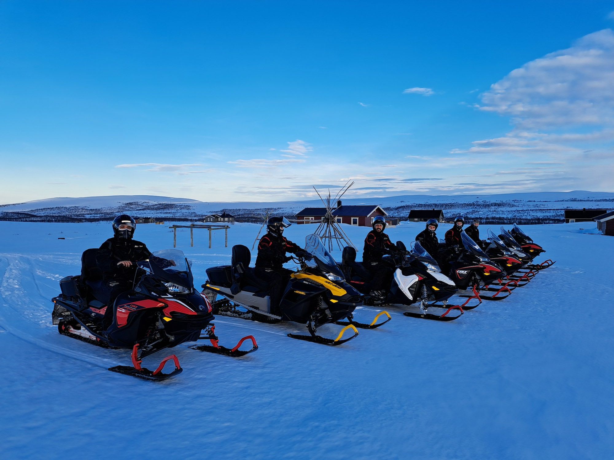 Snowmobilers line up in the Arctic wilderness, a Sami tent in the background.