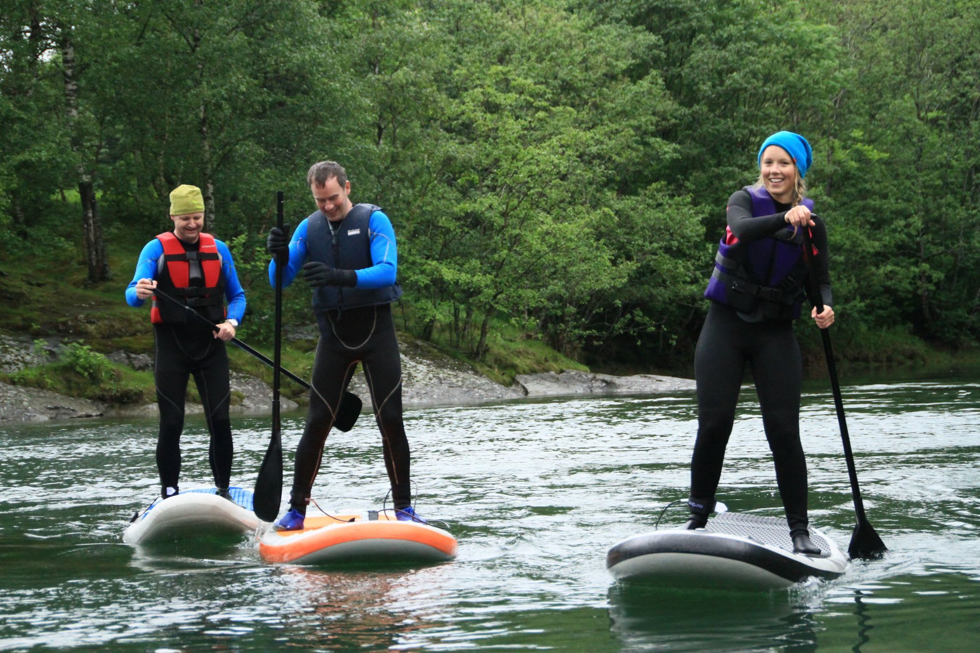 Three paddleboarders on a calm river, one smiling in a blue beanie, surrounded by greenery.