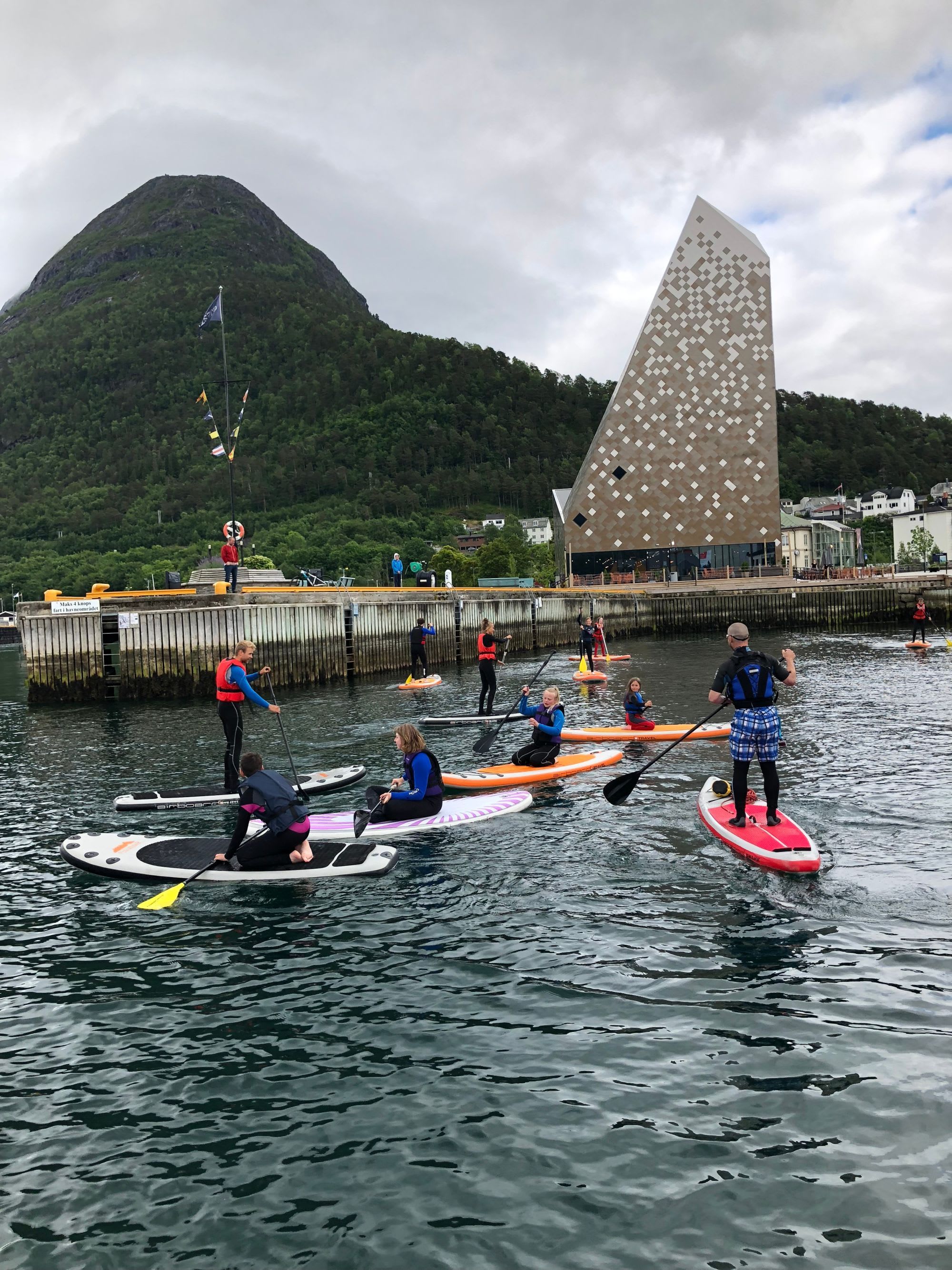 People paddleboarding near a harbor, with a modern building in the background and lush mountains surrounding.